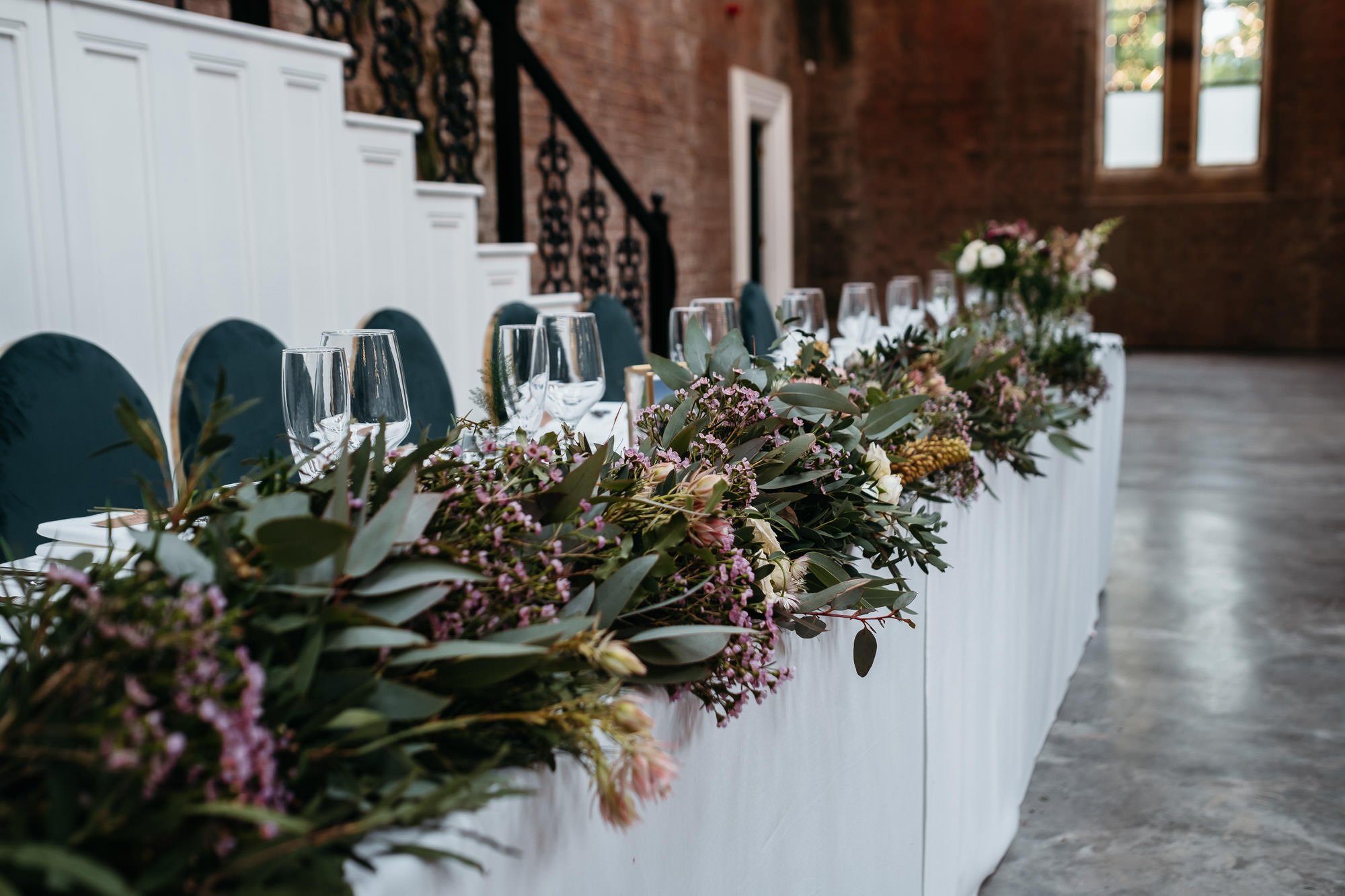 A long table set for a formal event with a floral centerpiece and empty wine glasses, in a room with brick walls and windows.