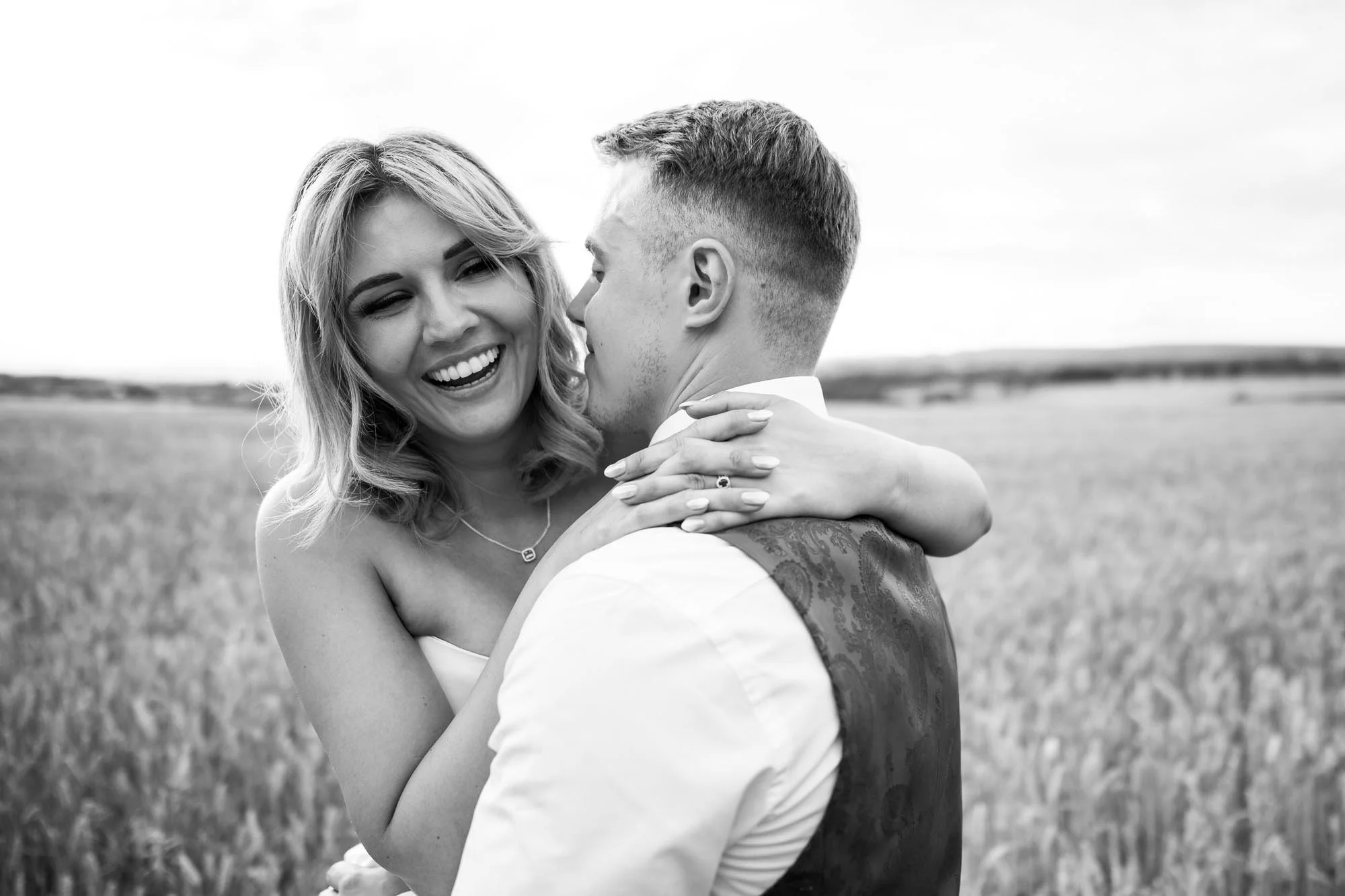 A black and white photograph of a joyful couple embracing outdoors in a field, with the woman smiling widely and the man close to her face, both appearing happy and affectionate.