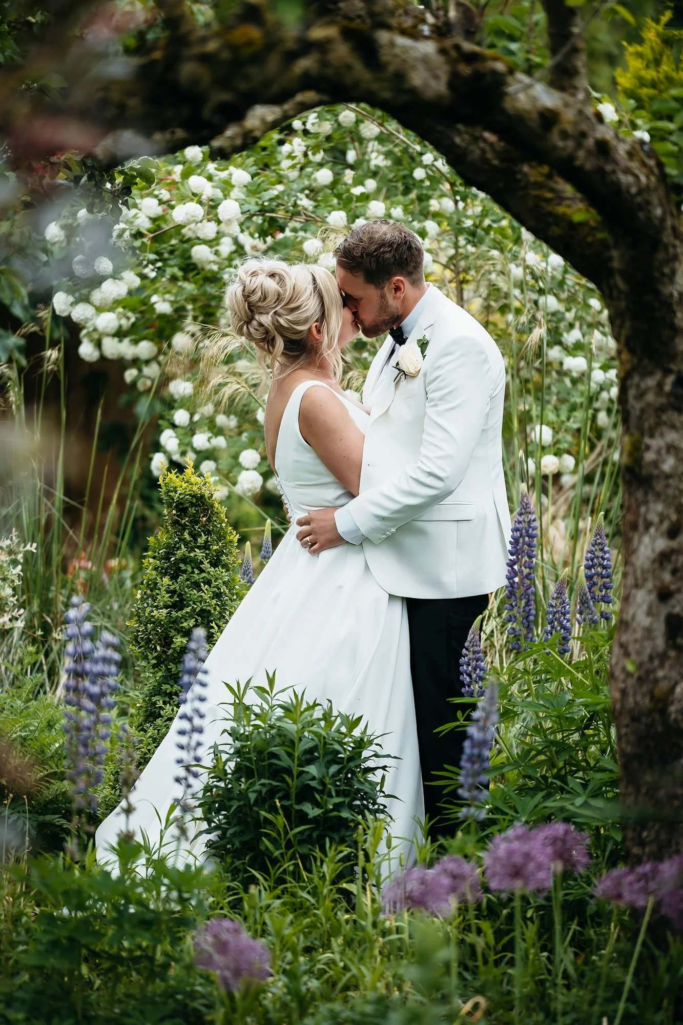 A bride and groom standing close together, kissing in a lush, green garden surrounded by white and purple flowers.