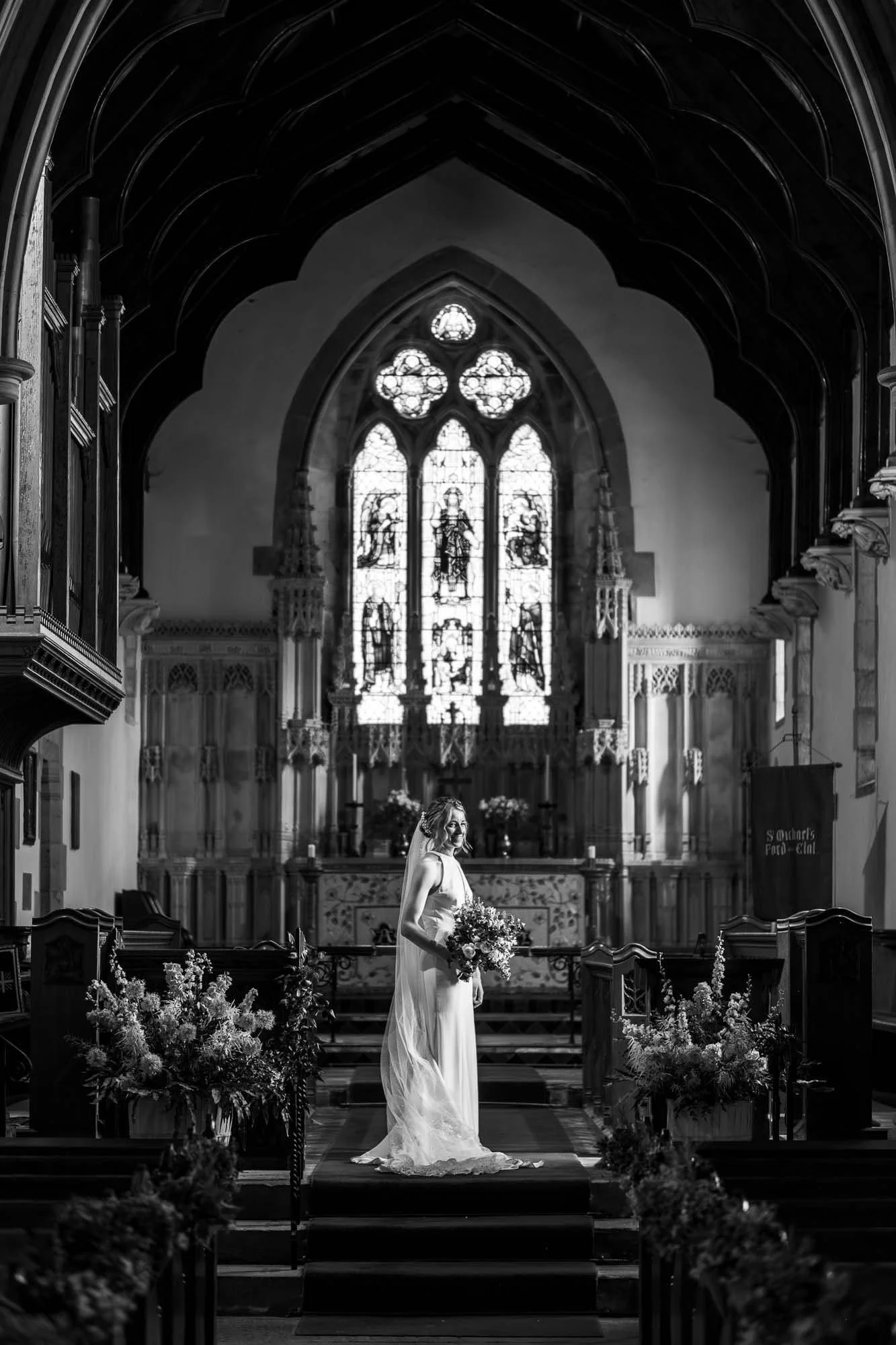 A bride in a white wedding dress holding a bouquet standing in a church with stained glass windows and gothic architecture.