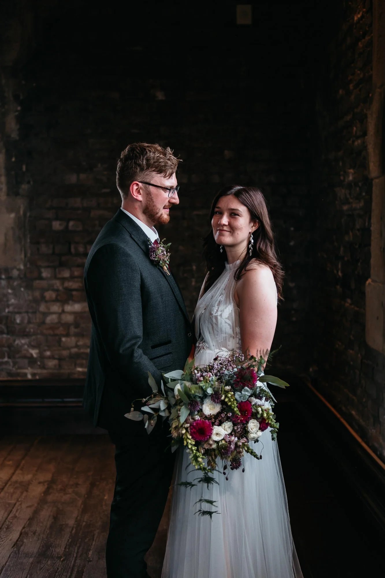 A bride and groom standing together indoors with a dark brick wall in the background. The bride is holding a large bouquet of flowers and wearing a white gown, while the groom is dressed in a dark suit and glasses.