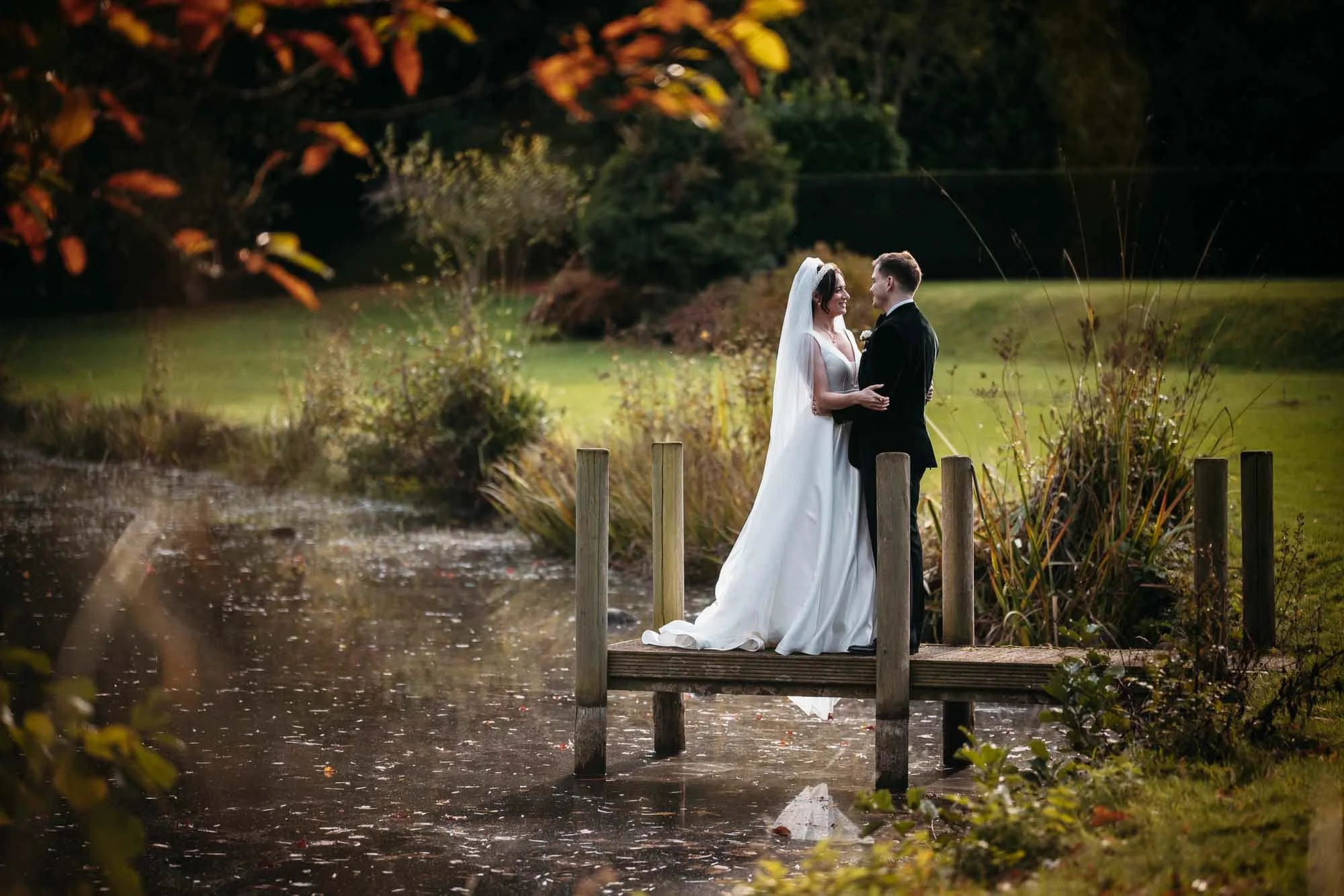 A bride and groom standing close together on a small dock over a pond, smiling and looking at each other, surrounded by autumn trees and foliage.