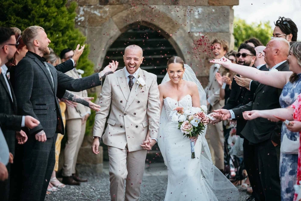 Bride and groom laughing beneath confetti at the stone archway at Danby Castle