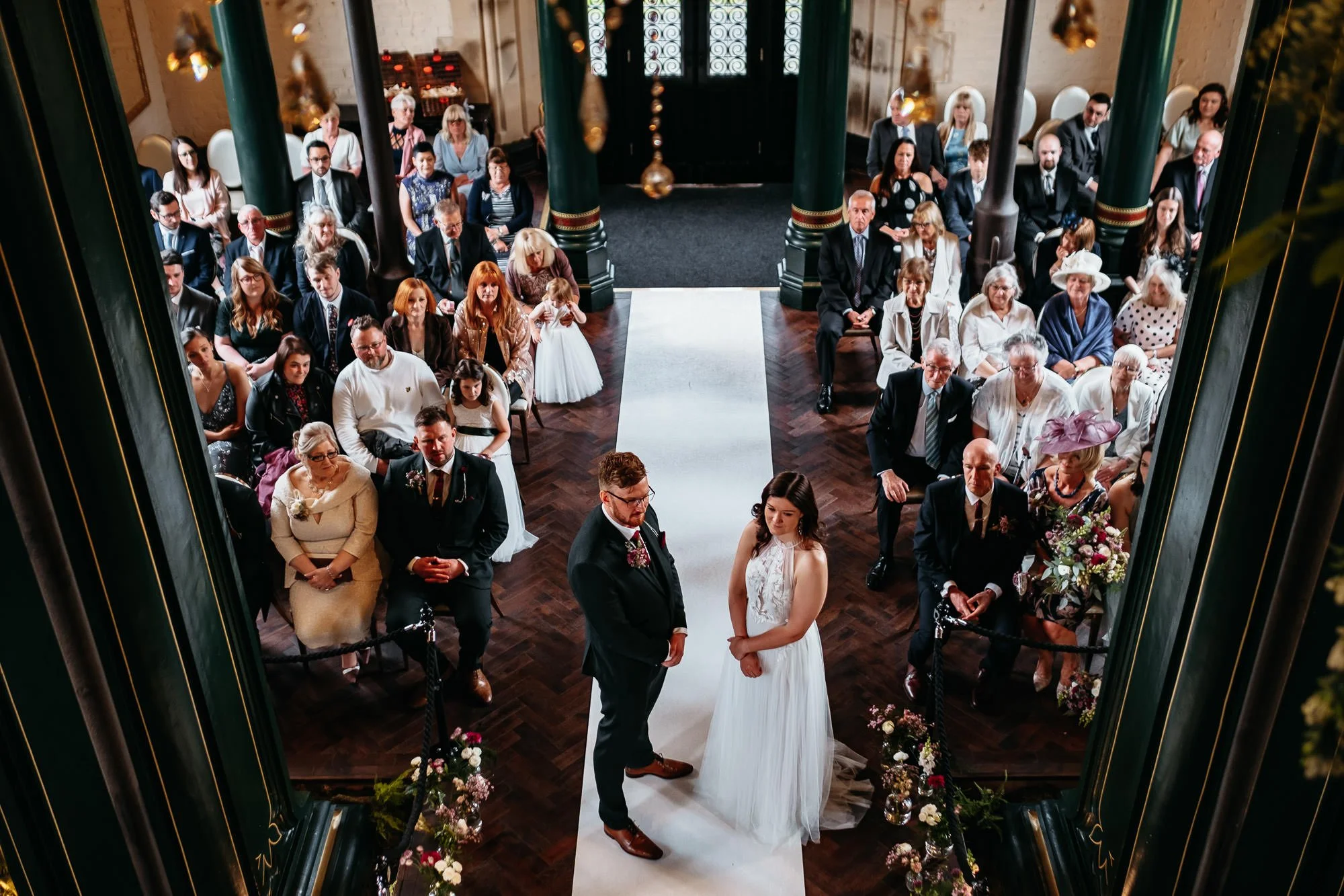A bride and groom standing at the altar during their wedding ceremony, surrounded by seated guests in a decorated indoor venue.
