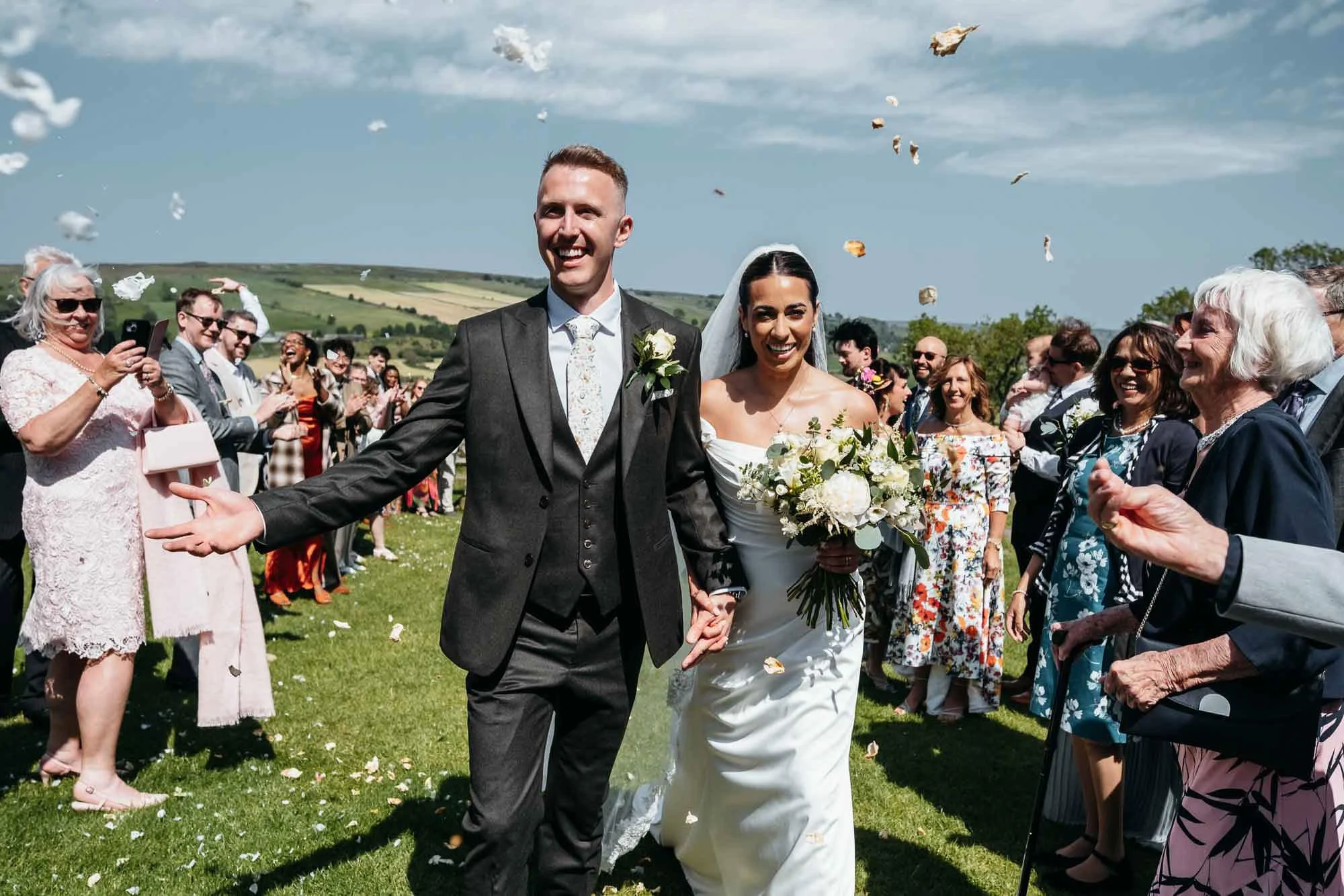 A bride and groom walking hand in hand through a line of guests throwing flower petals during their outdoor wedding ceremony on a sunny day with a green landscape and hills in the background.