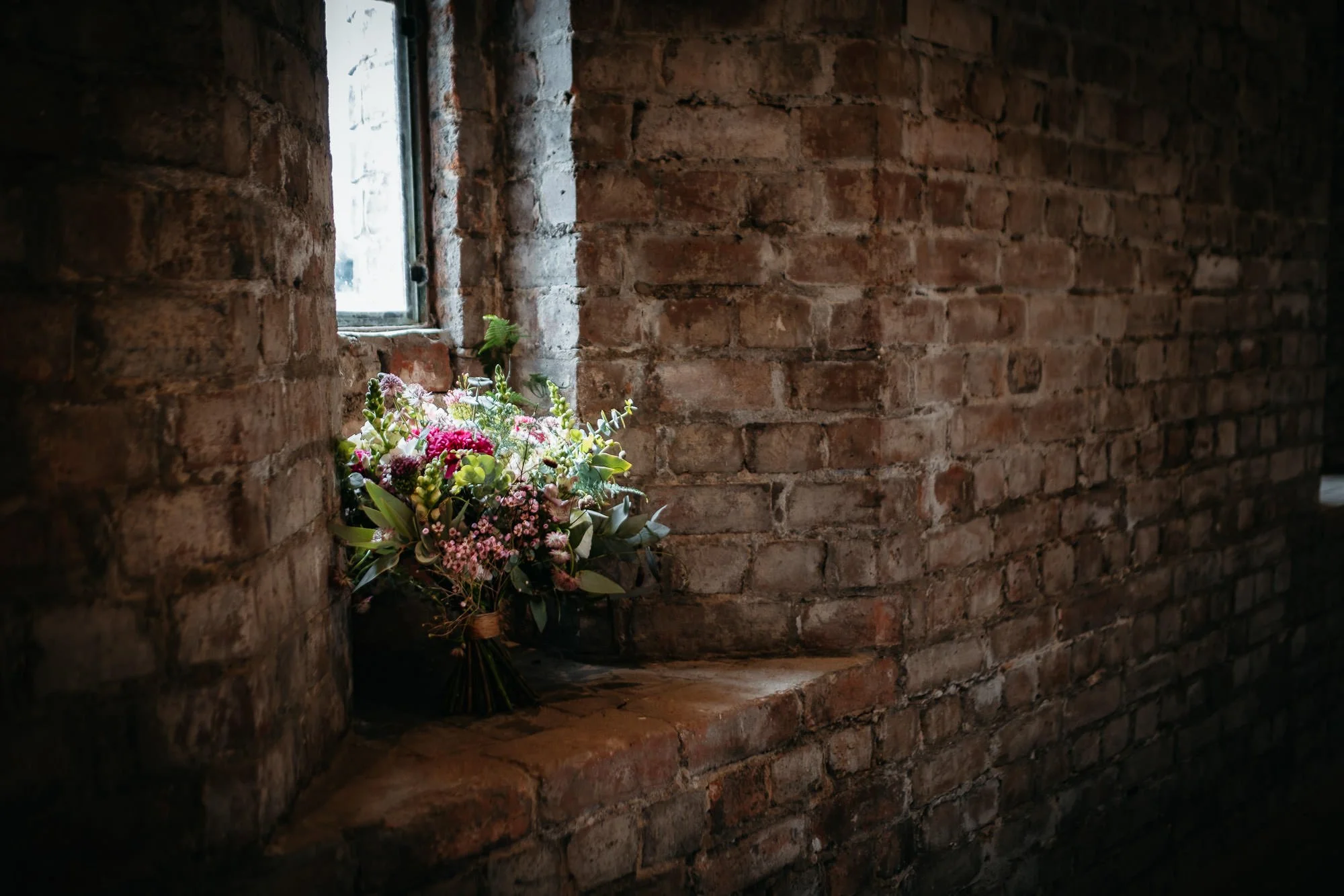 A bouquet of pink, white, and purple flowers resting on a brick window sill near a small window in a brick wall, with natural light coming through.