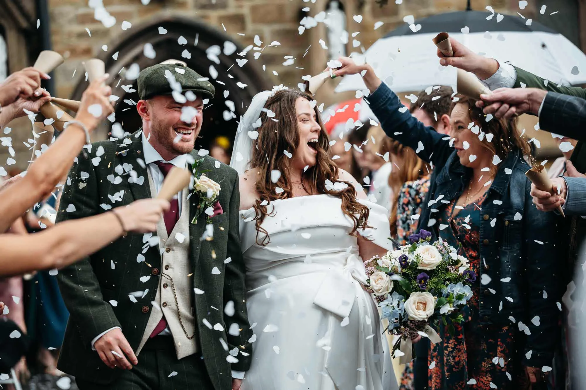 A bride and groom celebrating with guests, throwing white heart-shaped confetti outside a building, with a brick wall background. The bride is smiling and holding a bouquet.