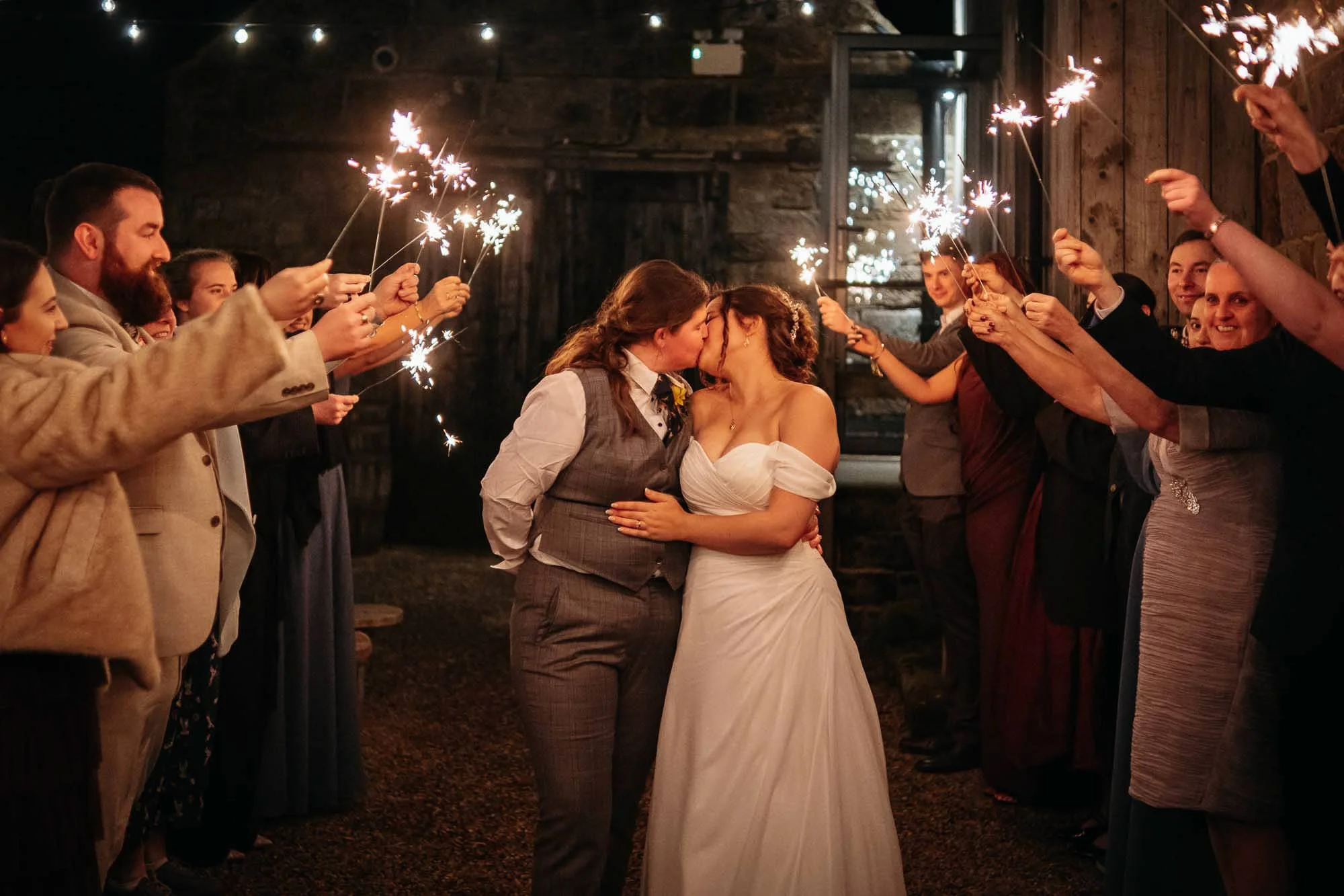 Two women in wedding attire sharing a kiss surrounded by guests holding sparklers at night.