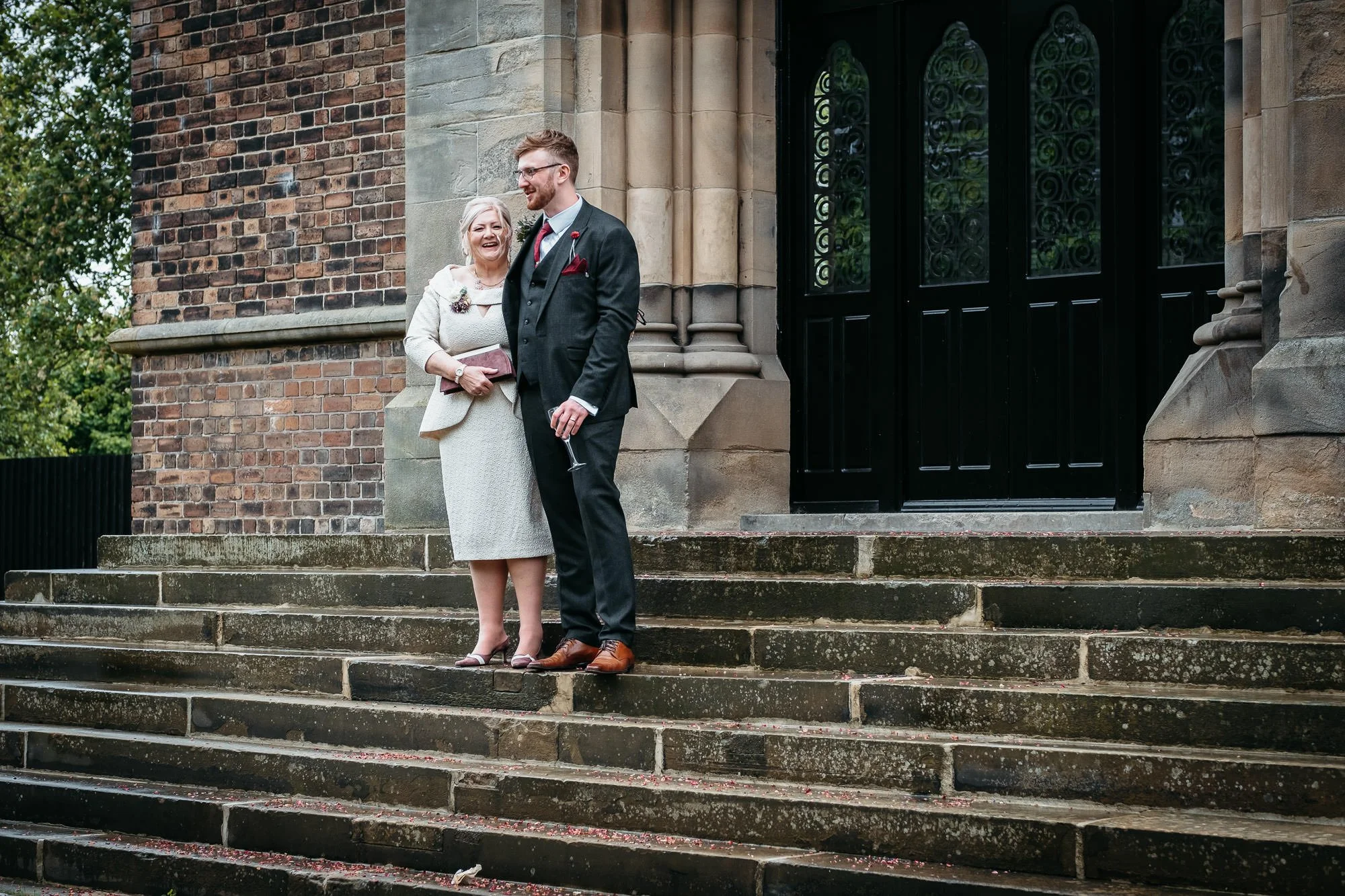 A joyful older woman and a smiling younger man in formal attire standing on church steps outside, with the woman holding a clutch and the man holding a glass. The background features a brick and stone building with an ornate black door.