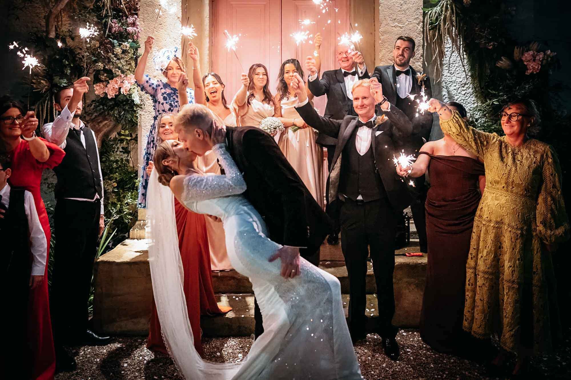 Bride and groom kissing as guests hold sparklers at the uplit pink door of The Croft Hotel at night