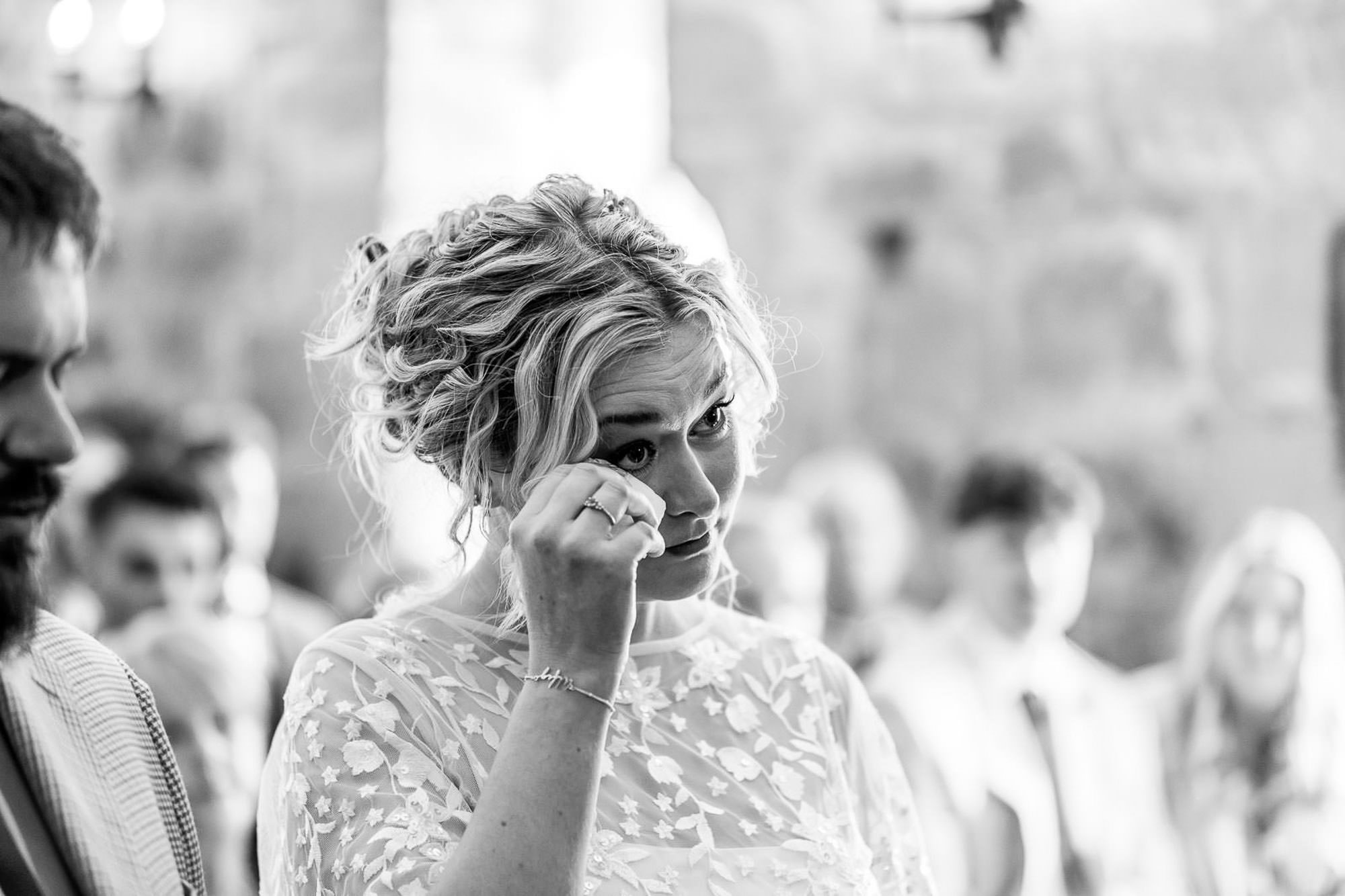 A woman with curly blonde hair wiping tears from her eye during a gathering, with blurred people in the background.