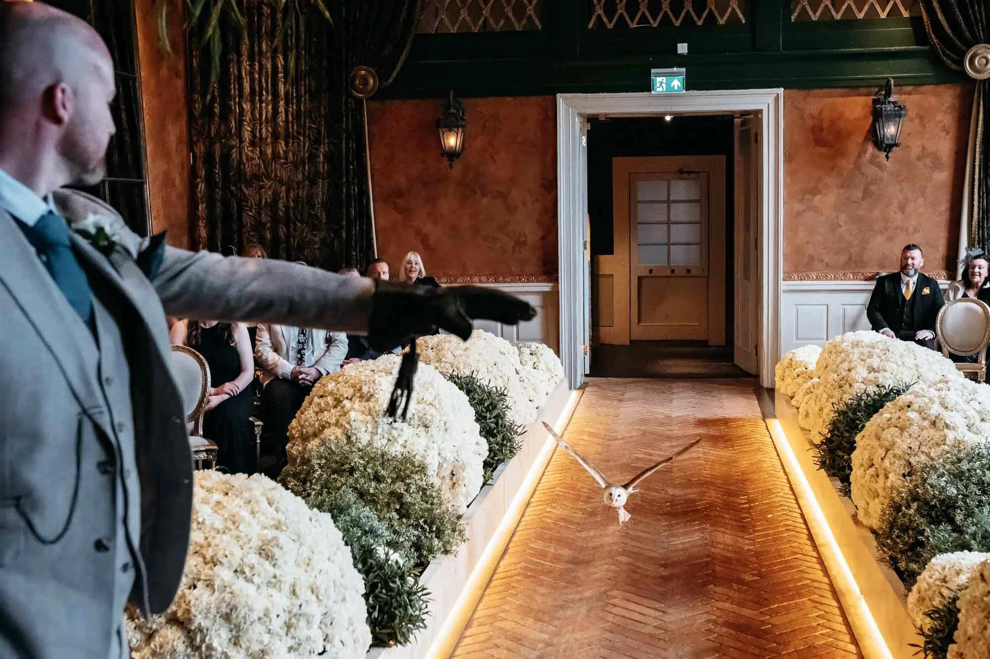 A wedding ceremony with a person holding a gloved sword or dagger, standing on an aisle decorated with white flowers. Guests are seated on either side, and there is a dog in the aisle. The room has ornate decor with wall lanterns, curtains, and an ex