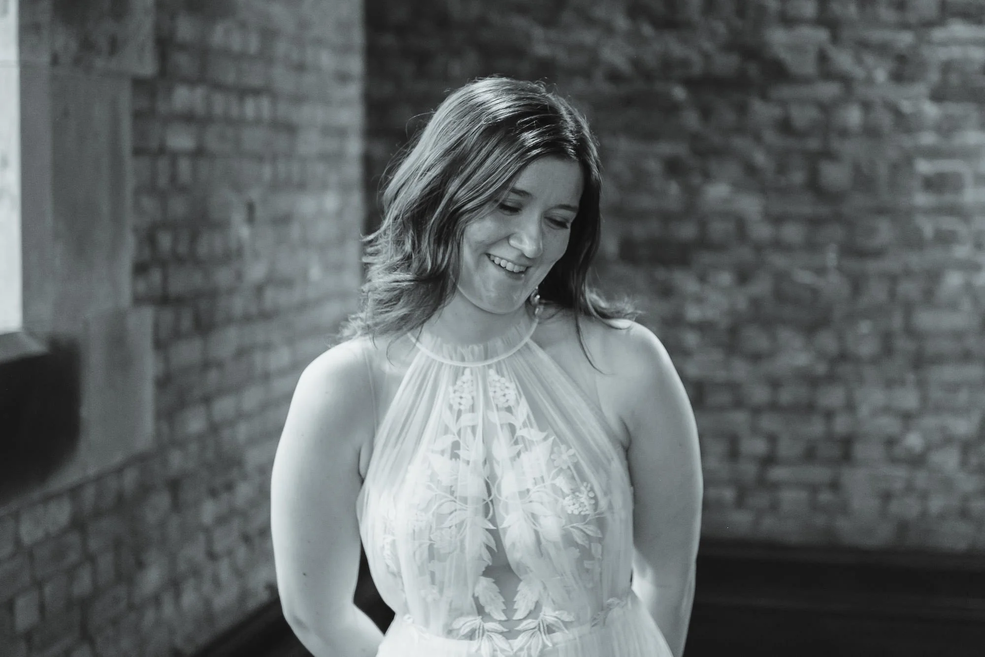 A woman with shoulder-length wavy hair is smiling and looking downward. She is wearing a sleeveless, lace dress with a floral pattern. The background features a brick wall.