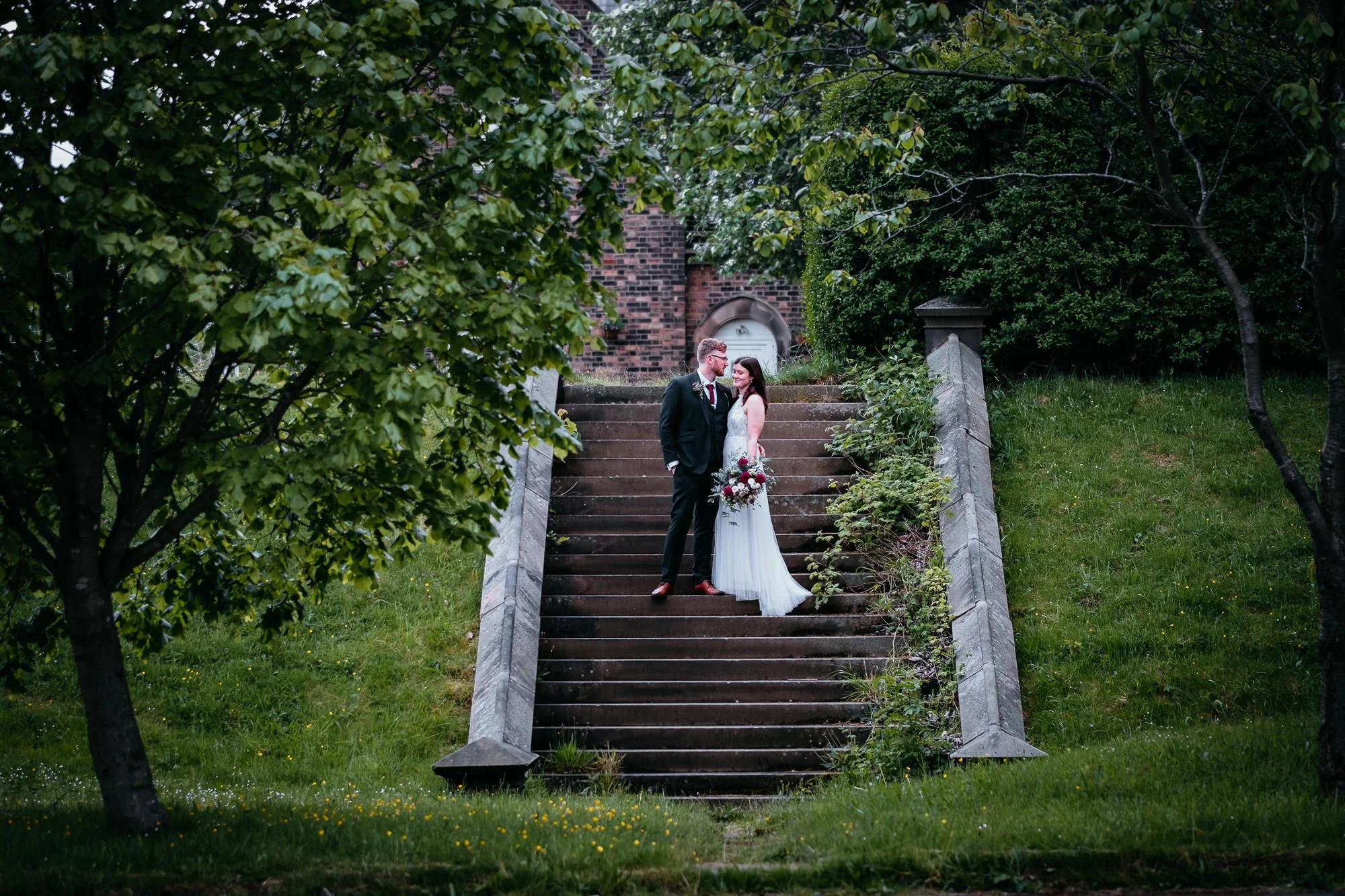 A bride and groom standing on outdoor steps, surrounded by greenery, during their wedding. The bride is holding a bouquet, and they are looking at each other.
