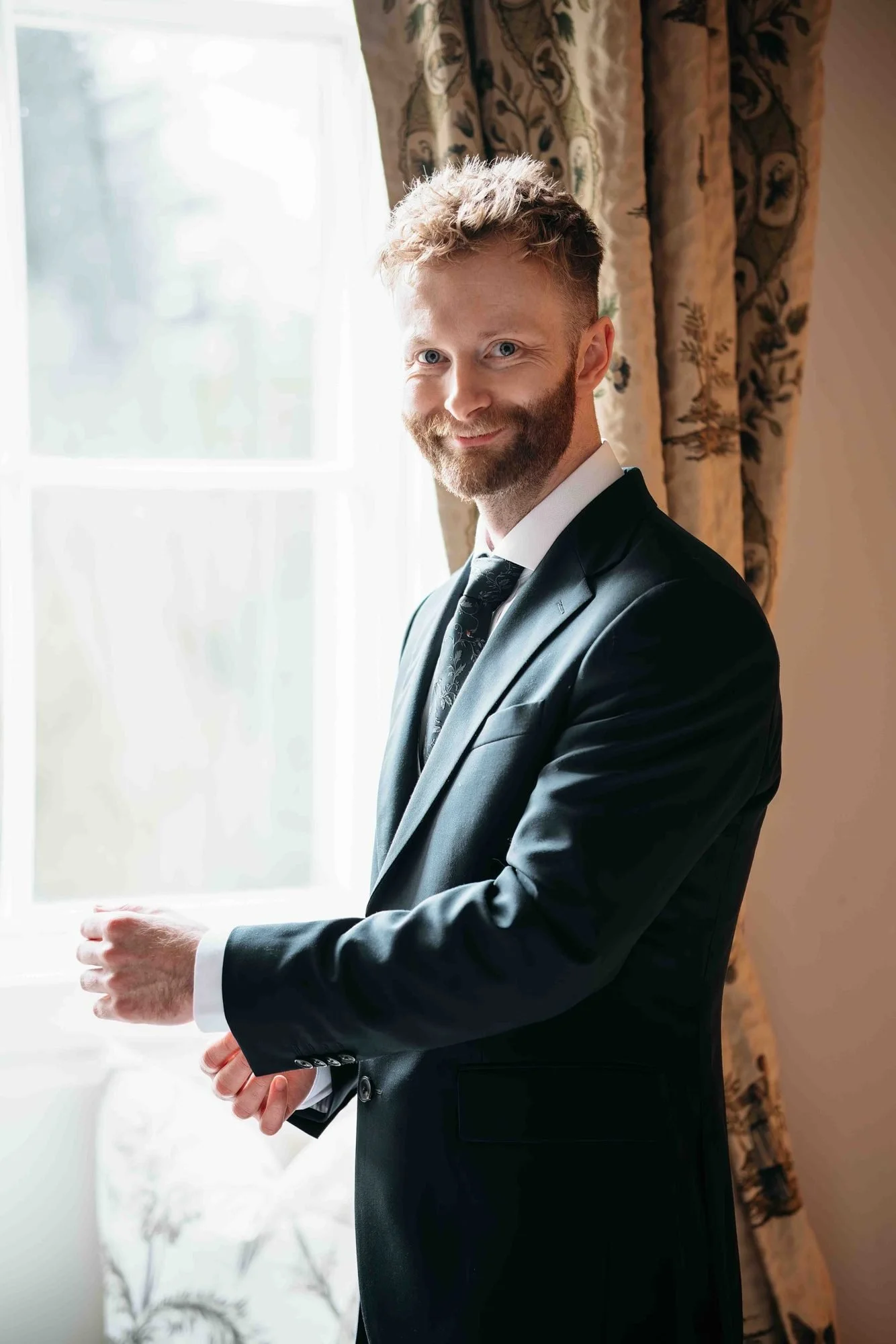 Groom smiling by a tall sash window while getting ready at Middleton Lodge