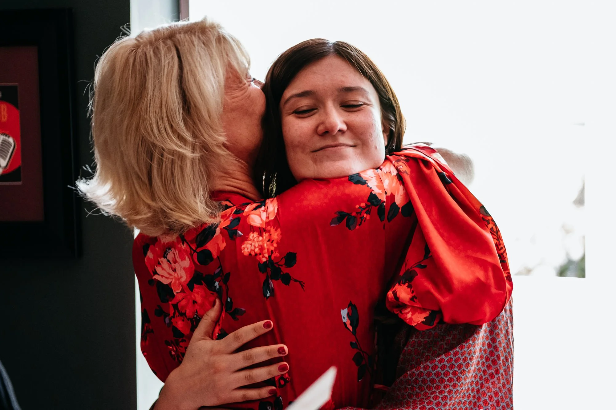 An older woman with blonde hair giving a hug and kiss on the cheek to a young woman with dark hair, both wearing red floral clothing, inside a room with a bright background.