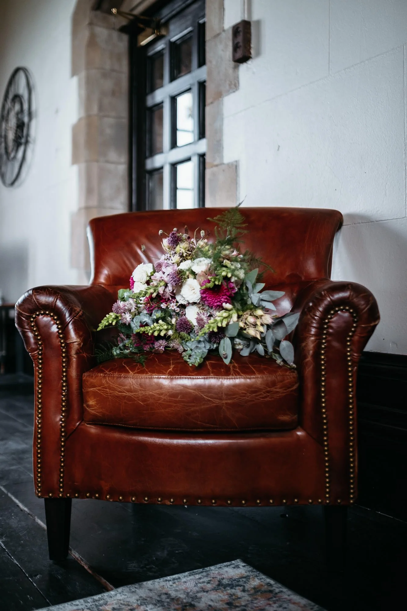 A brown leather armchair with a bouquet of flowers on it, positioned near a window with a black frame inside a room with stone walls.