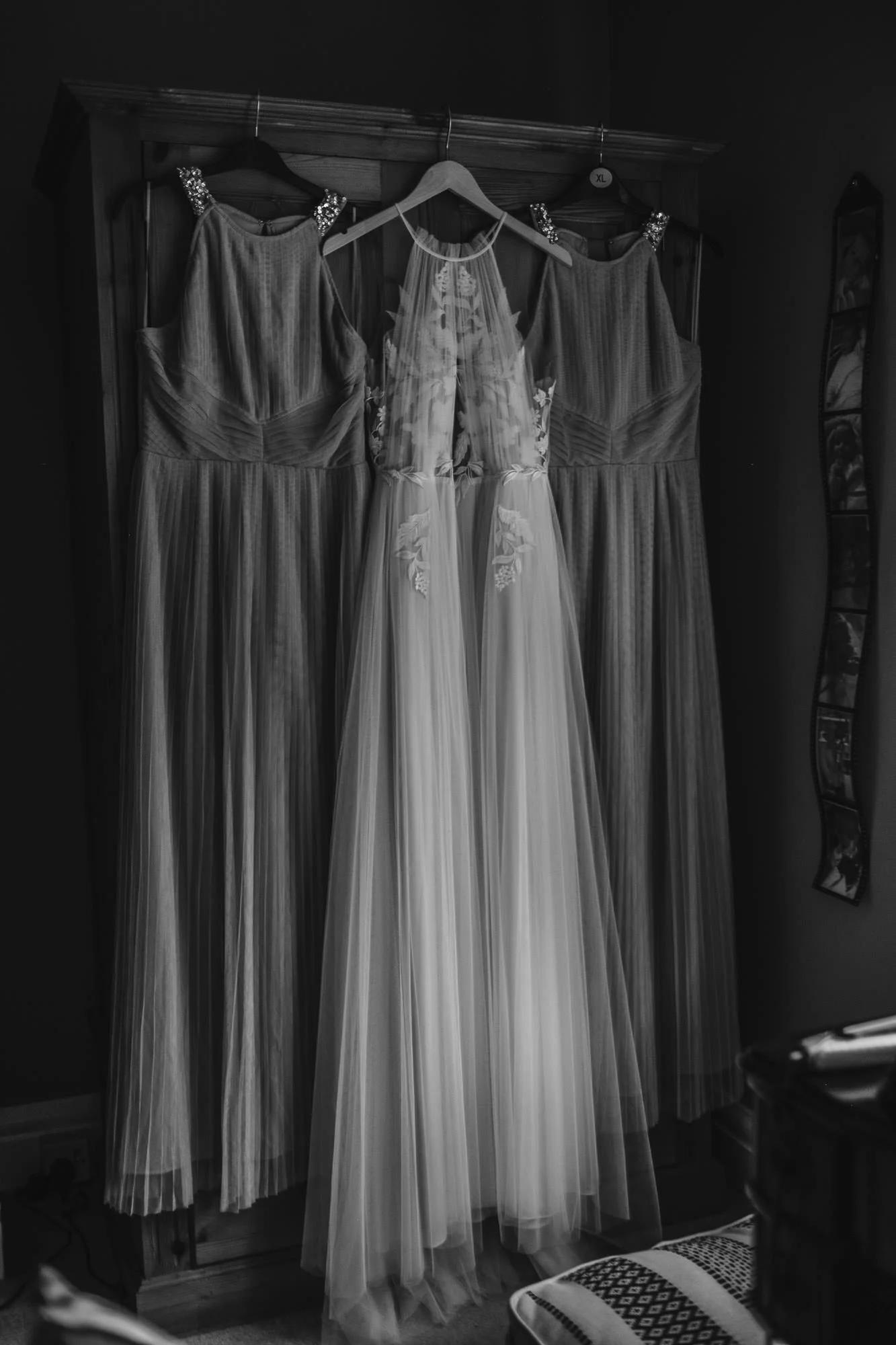 Three dresses hanging on a wooden wardrobe, including a wedding dress with floral embroidery in the center and two matching bridesmaid dresses on either side.
