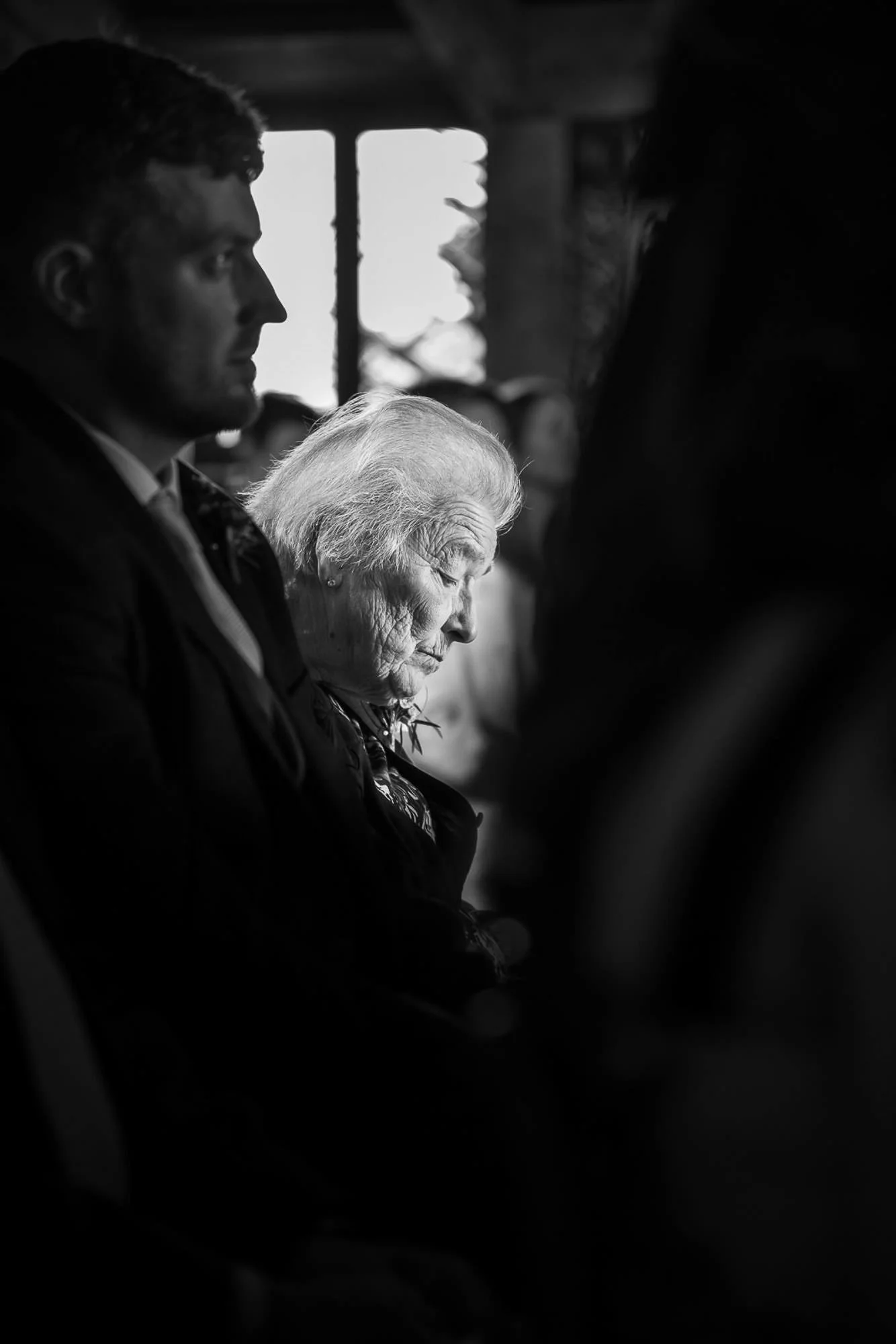 A black and white photo of a man and an elderly woman sitting side by side, both with serious expressions, inside a dimly lit space with a window in the background.