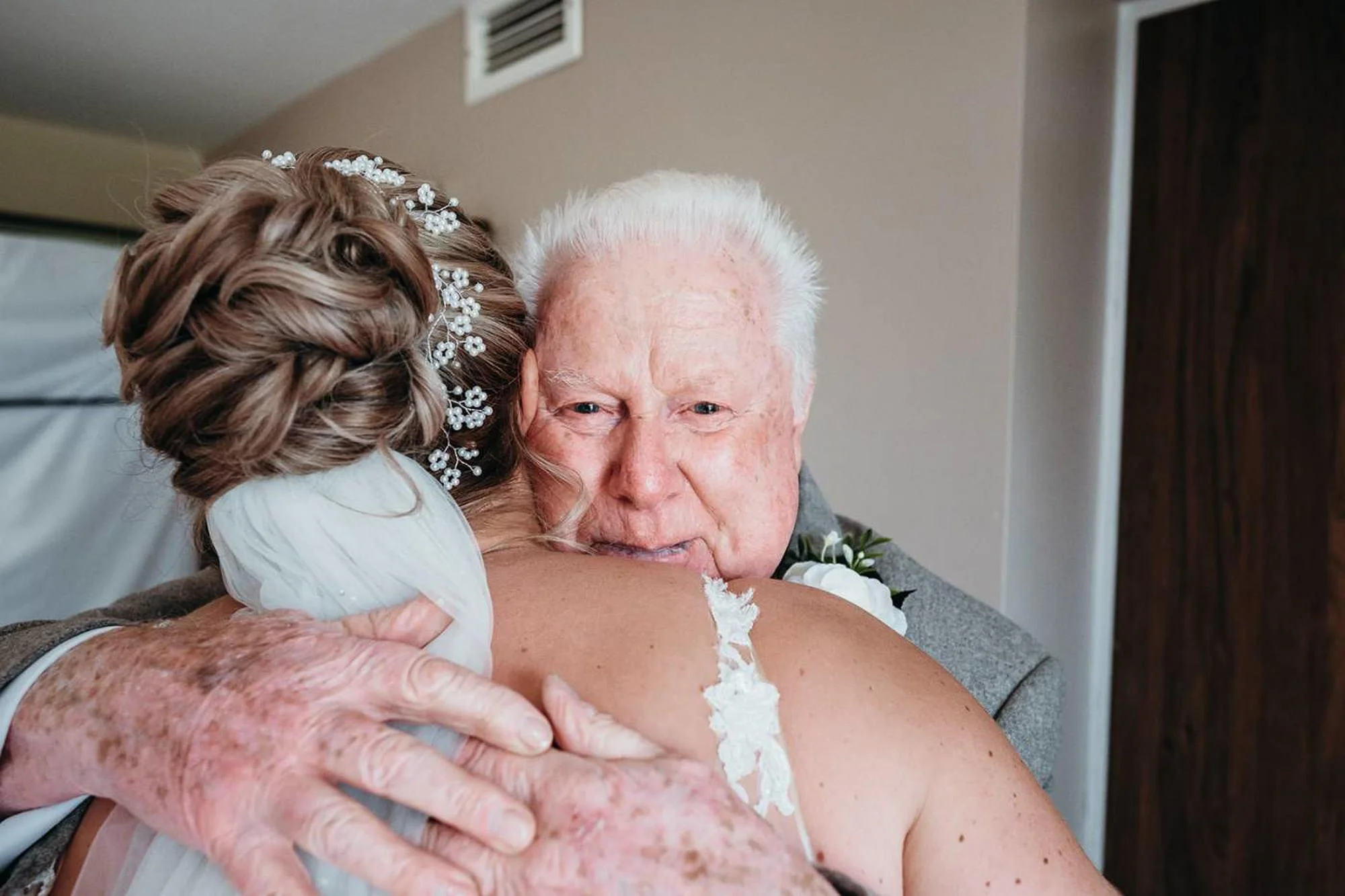An older woman and a woman with styled hair and a bead tiara are hugging in a room, possibly a hospital room, with the older woman smiling gently.