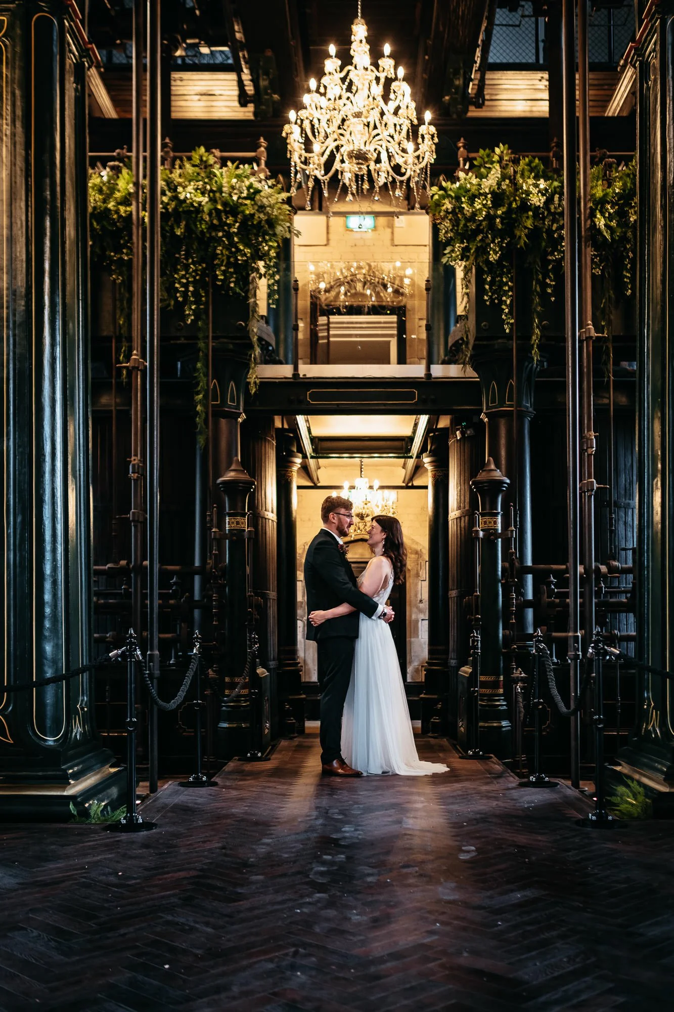 A bride and groom stand face to face, holding each other in a historic, elegant building with dark wood and ornate architectural details, illuminated by a large chandelier overhead.