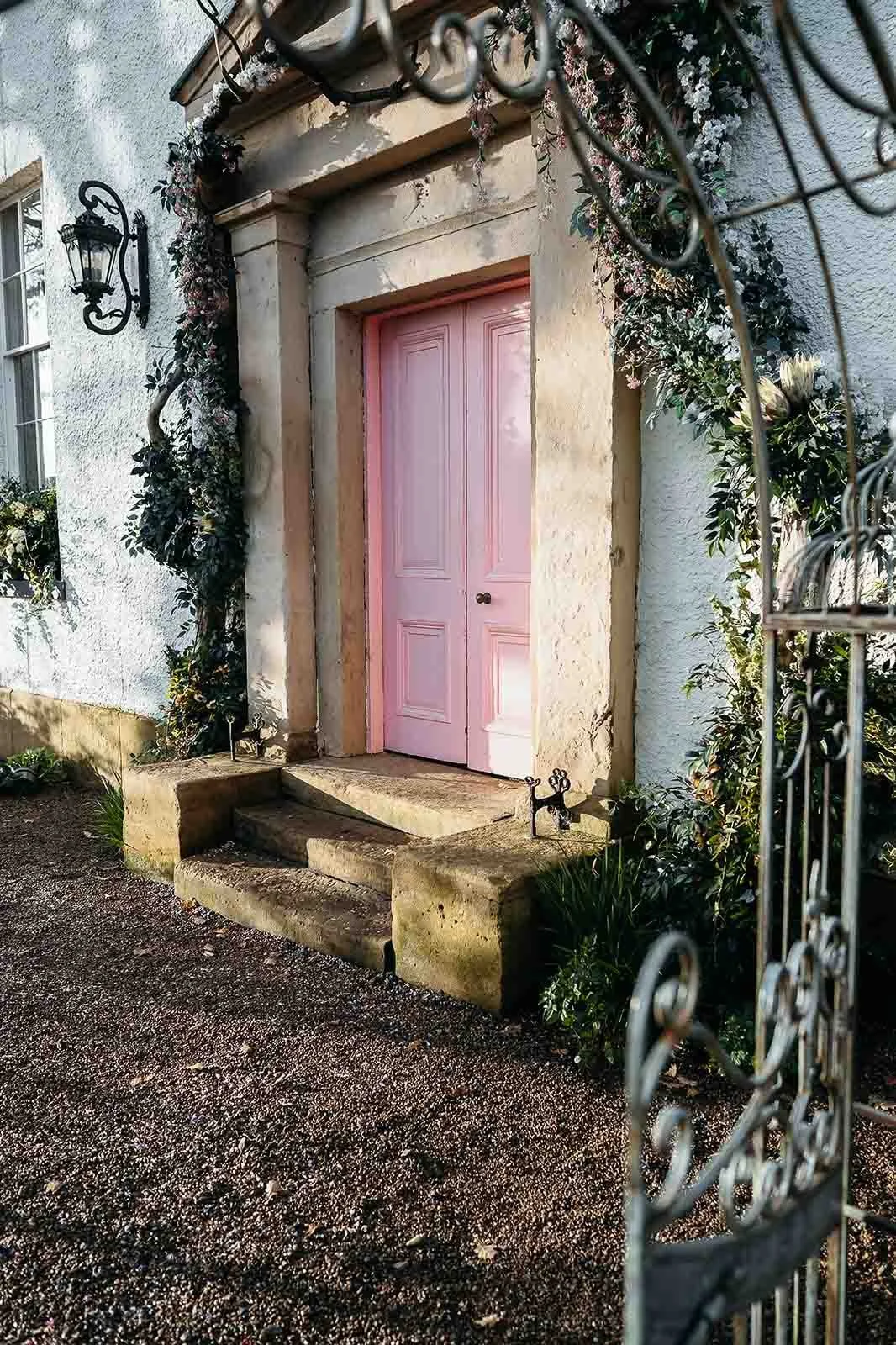 The pink door and stone steps at The Croft Hotel, framed by foliage and a wrought iron arch