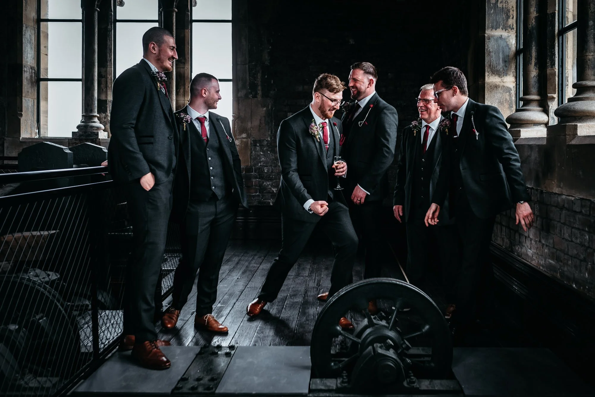 Group of seven men dressed in tuxedos with boutonnières, standing and laughing in a rustic, industrial-style room with brick walls and large windows.