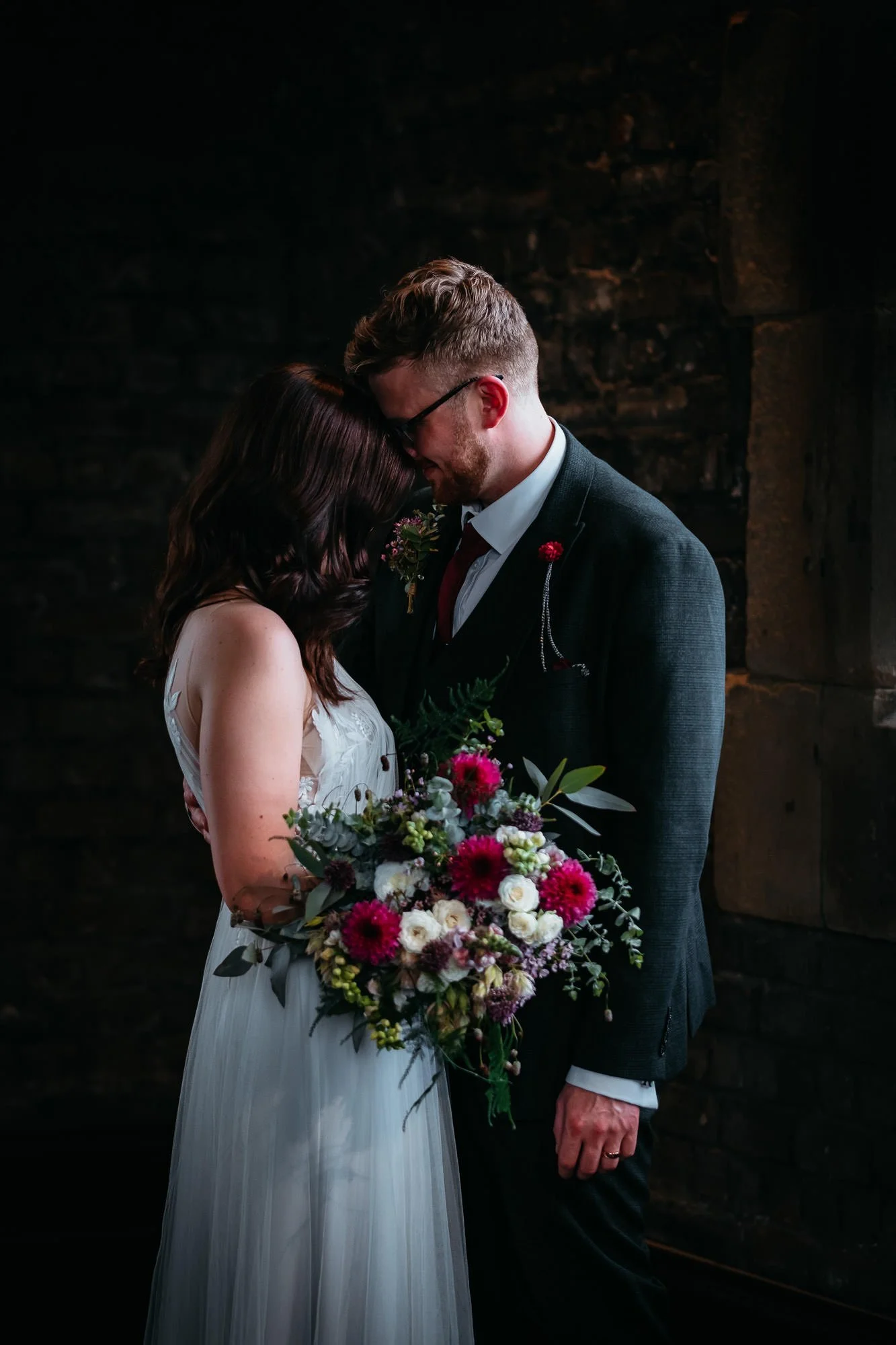 Wedding couple with foreheads touching, holding a bouquet of flowers in front of a dark brick wall.