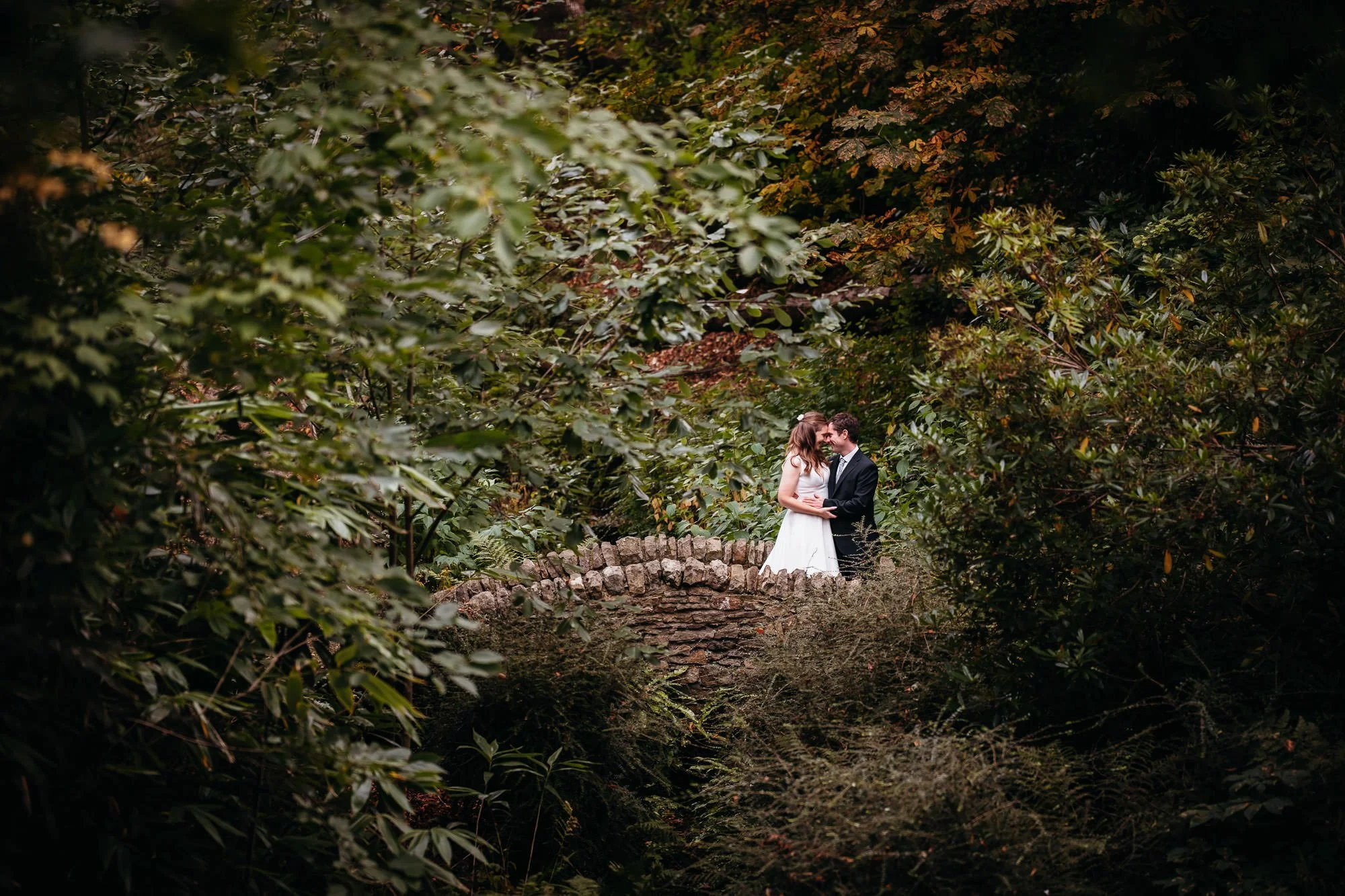 A bride and groom standing on a stone bridge in a forest, surrounded by dense green foliage, sharing a close and loving moment.