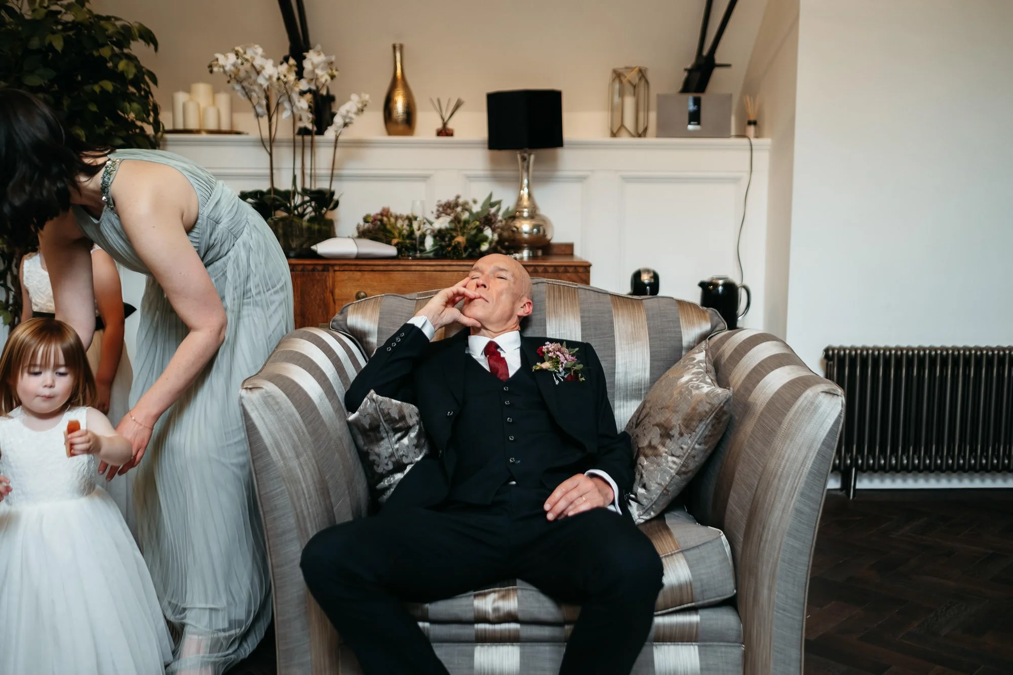 An elderly man in a black suit with a red tie and boutonniere is sitting on a striped sofa, yawning with his eyes closed, holding his face with one hand. A woman and a young girl in white dresses are nearby, with the woman leaning over towards the gi