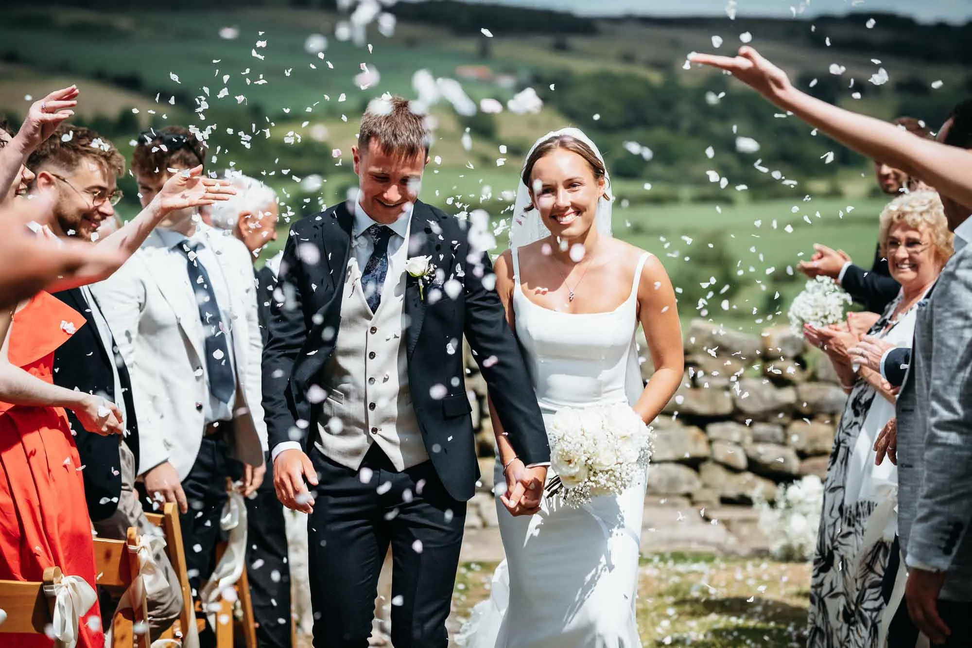 A bride and groom walking hand in hand outdoors after exchanging vows, surrounded by smiling guests tossing white confetti during a wedding celebration in a scenic green rural area.