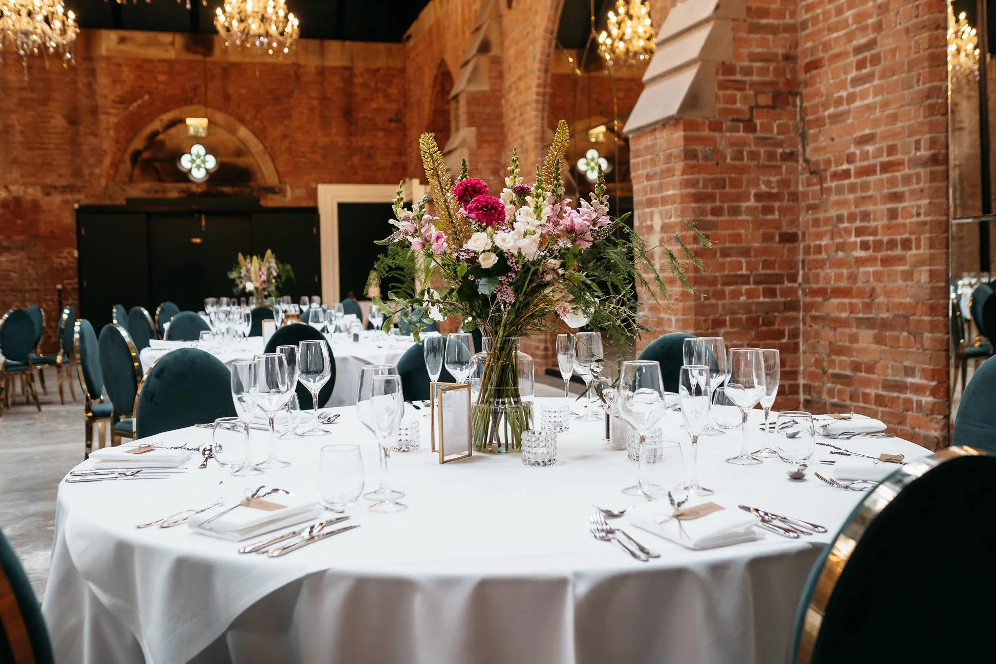 A wedding or banquet reception setup in an industrial-style venue with exposed brick walls and large arched windows, featuring a round table decorated with a tall floral centerpiece of pink, white, and purple flowers with greenery, surrounded by glas