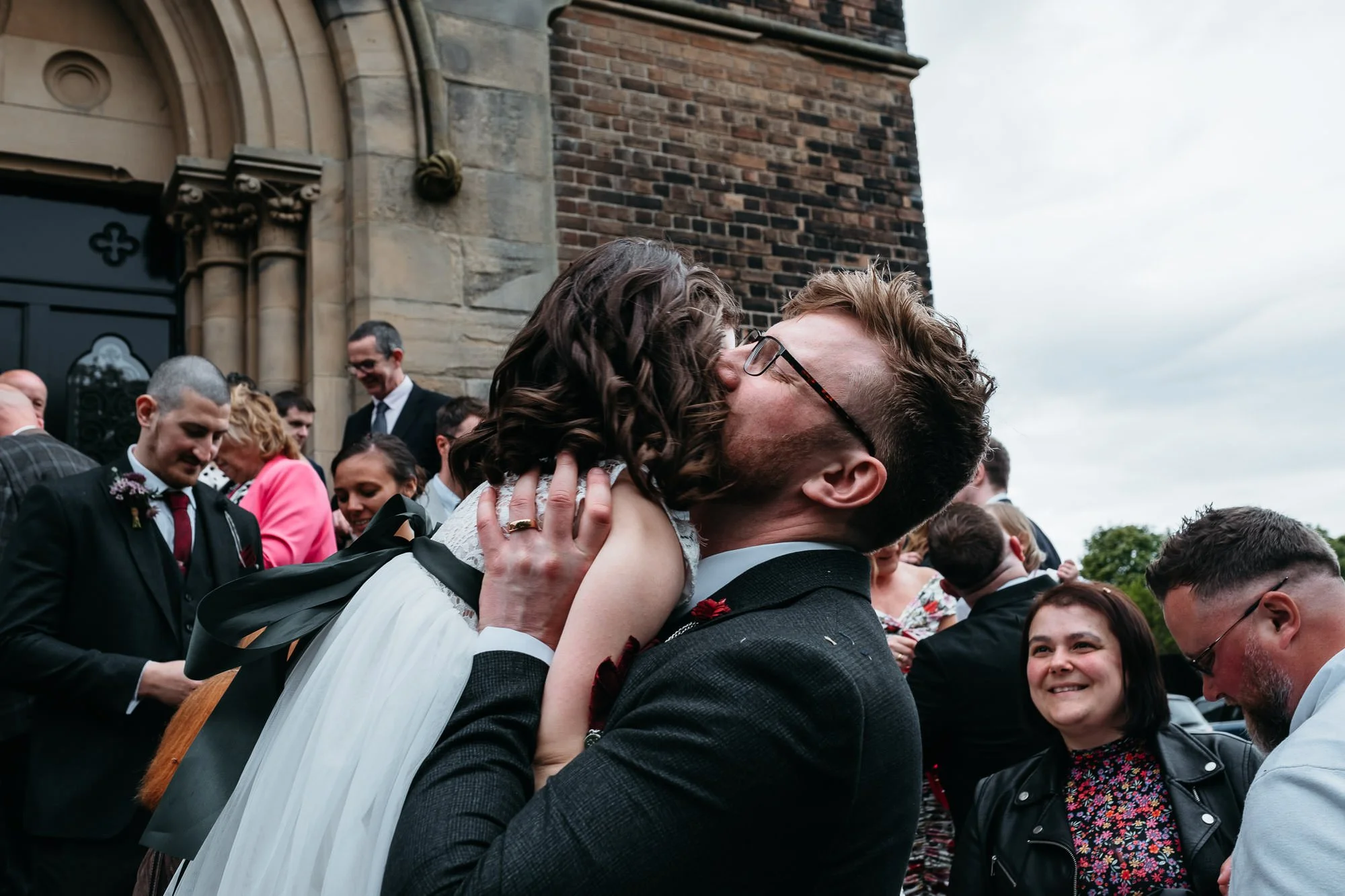 A man with glasses and a woman with dark, curly hair kissing, surrounded by people outside a church.