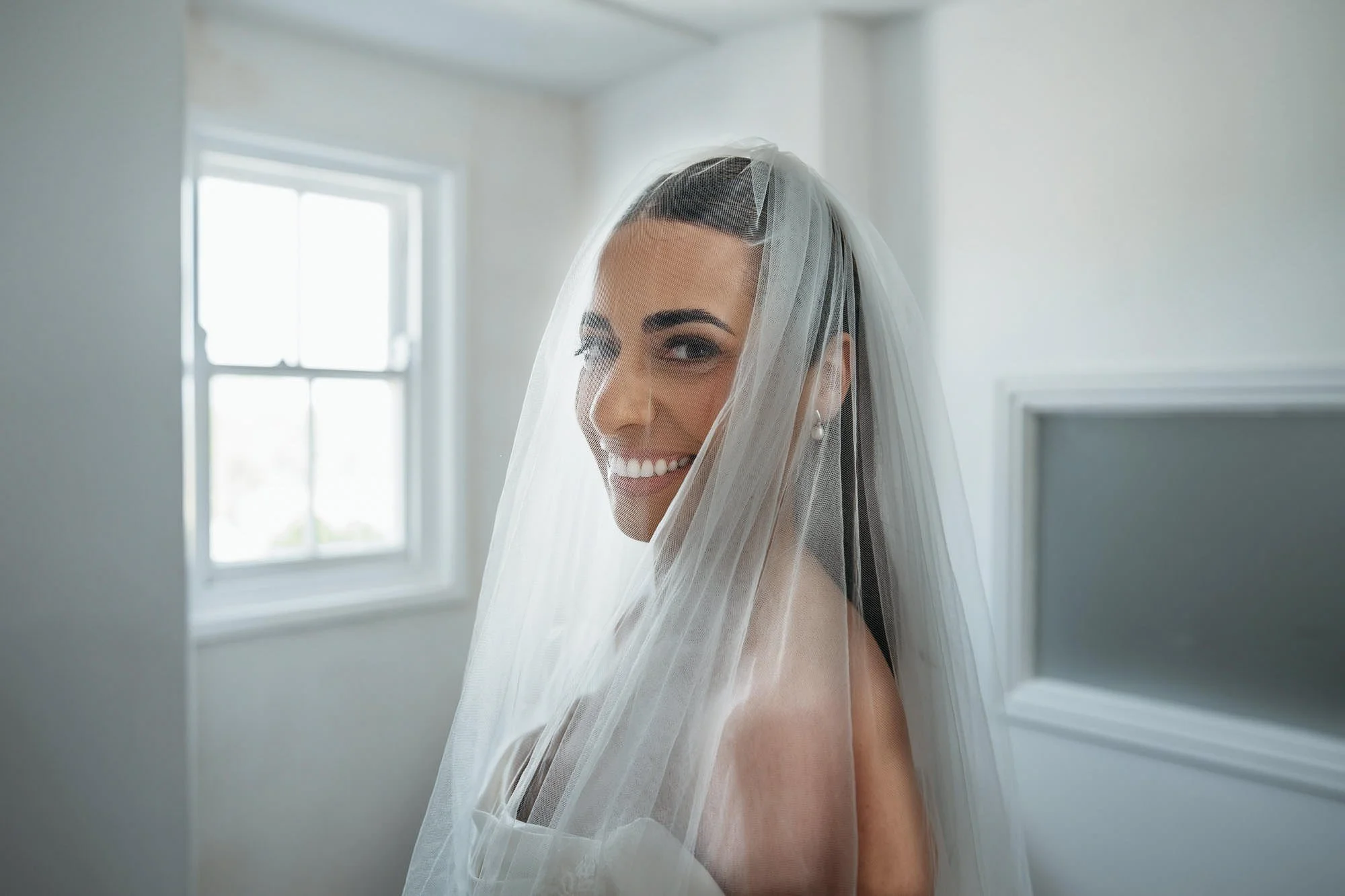 A smiling woman with dark hair and makeup, wearing a wedding veil and pearl earrings, is indoors near a window with white walls and a decorative wall feature.