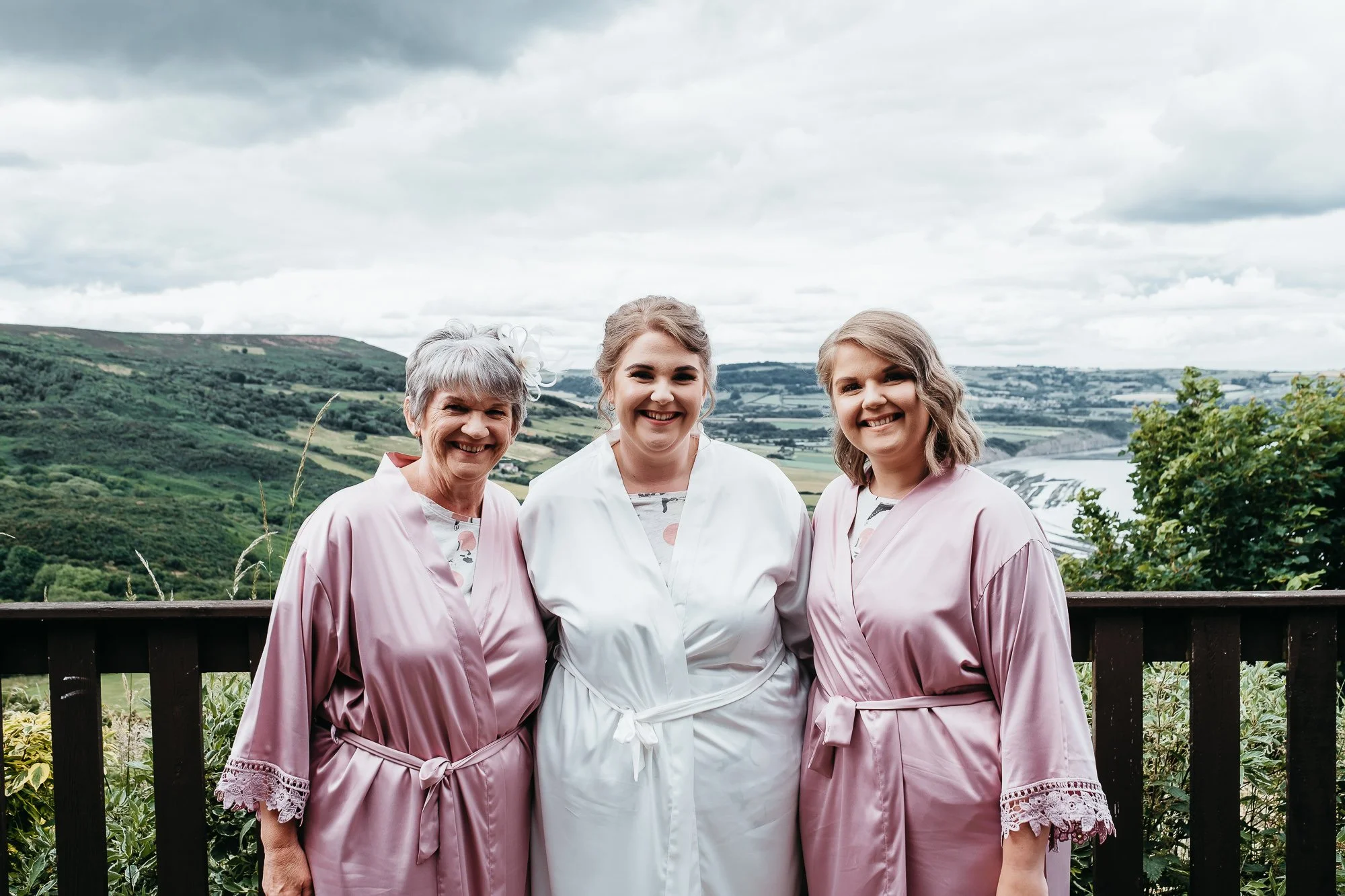 Bride with two women in pink on the balcony above the valley and coast at a Grand Villa Heights wedding, Ravenscar