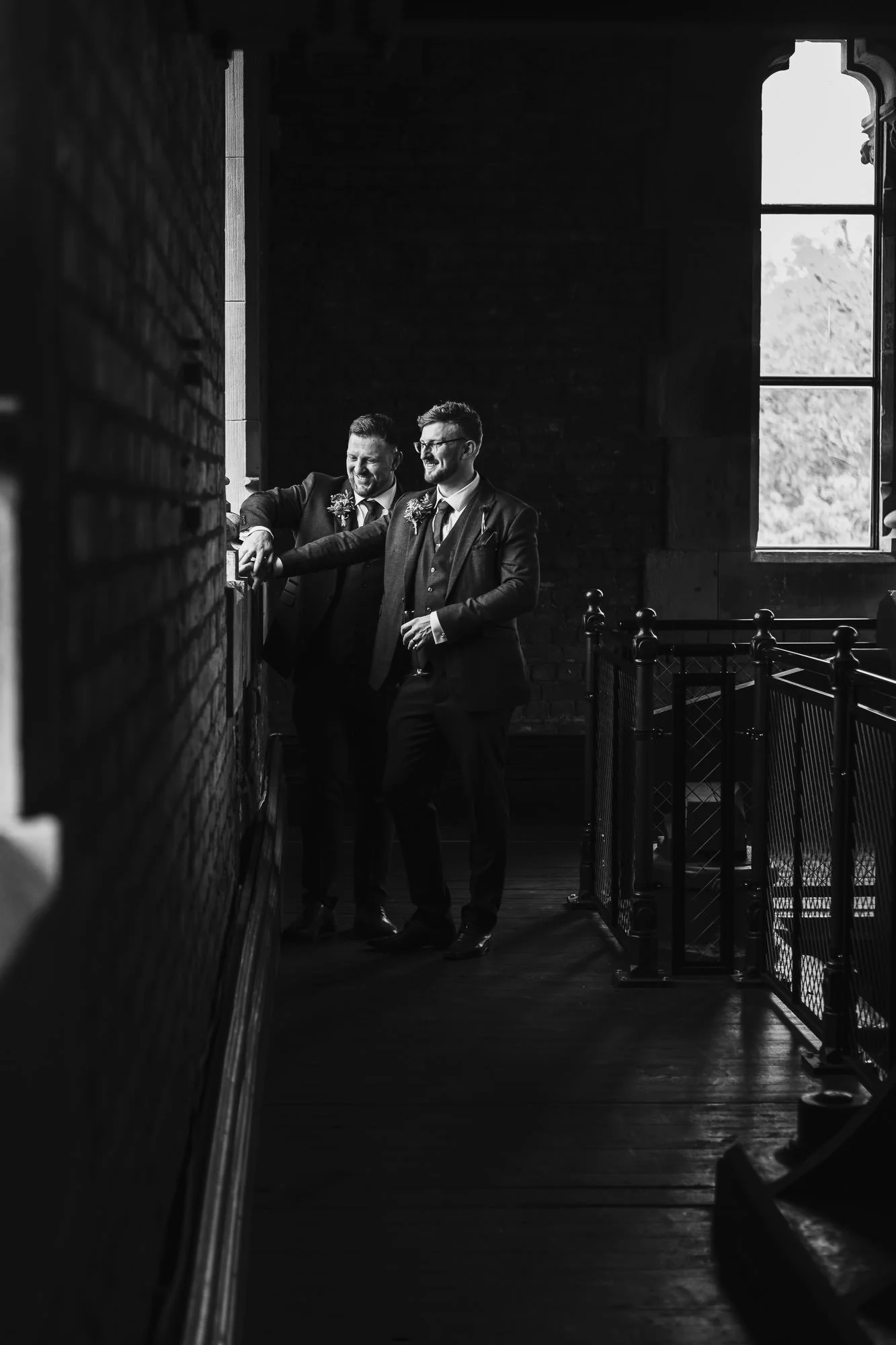 Two men in suits standing inside a building, smiling and talking near a brick wall and a window.