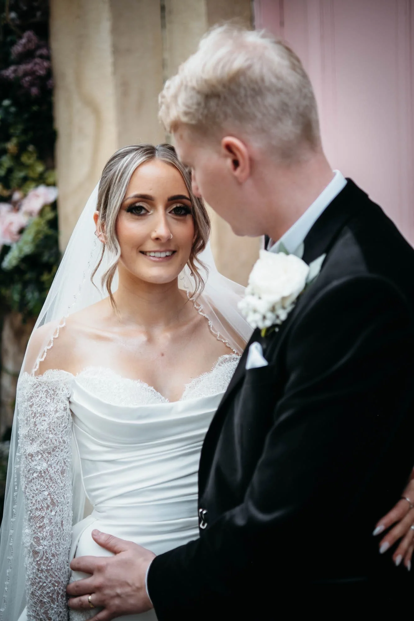 Bride in a white lace wedding dress and veil looking at groom, who is in a black tuxedo with a white shirt, holding her hand during a wedding ceremony. The groom has blond hair, and the bride has light brown hair styled in loose waves.