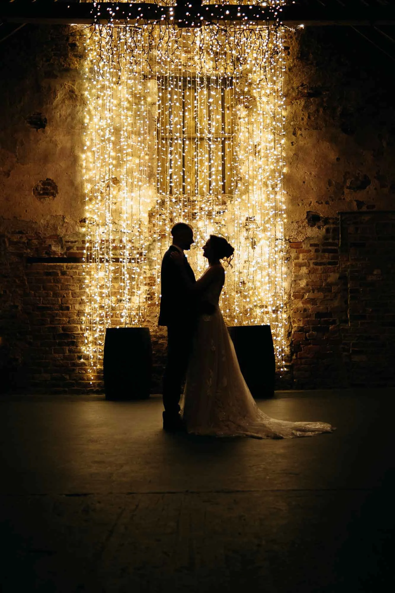 Silhouette of a bride and groom holding each other in front of a wall of hanging fairy lights with a brick wall background.