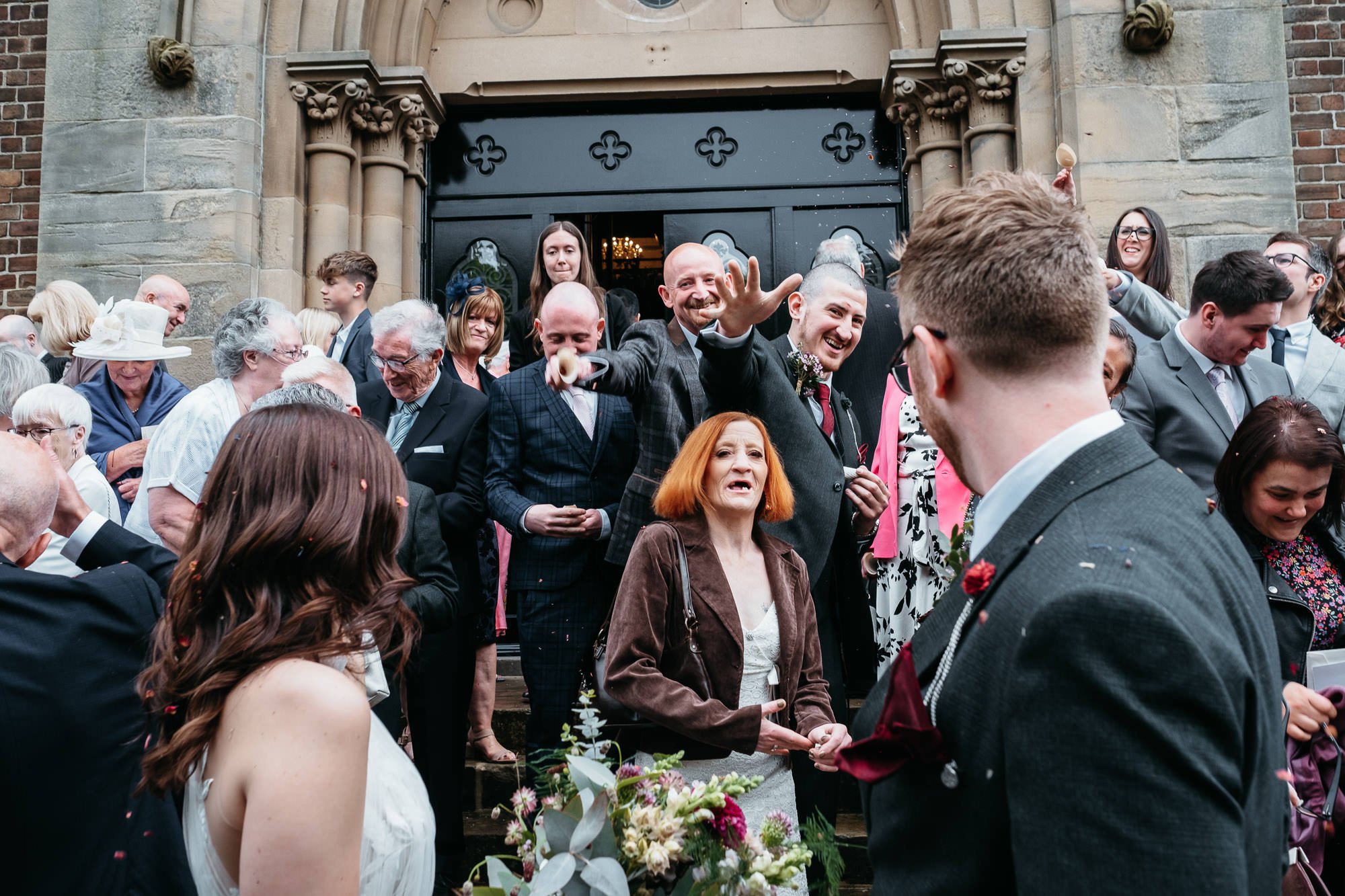 People gathered outside a church, celebrating, with some throwing confetti, and a woman holding a bouquet of flowers in front.