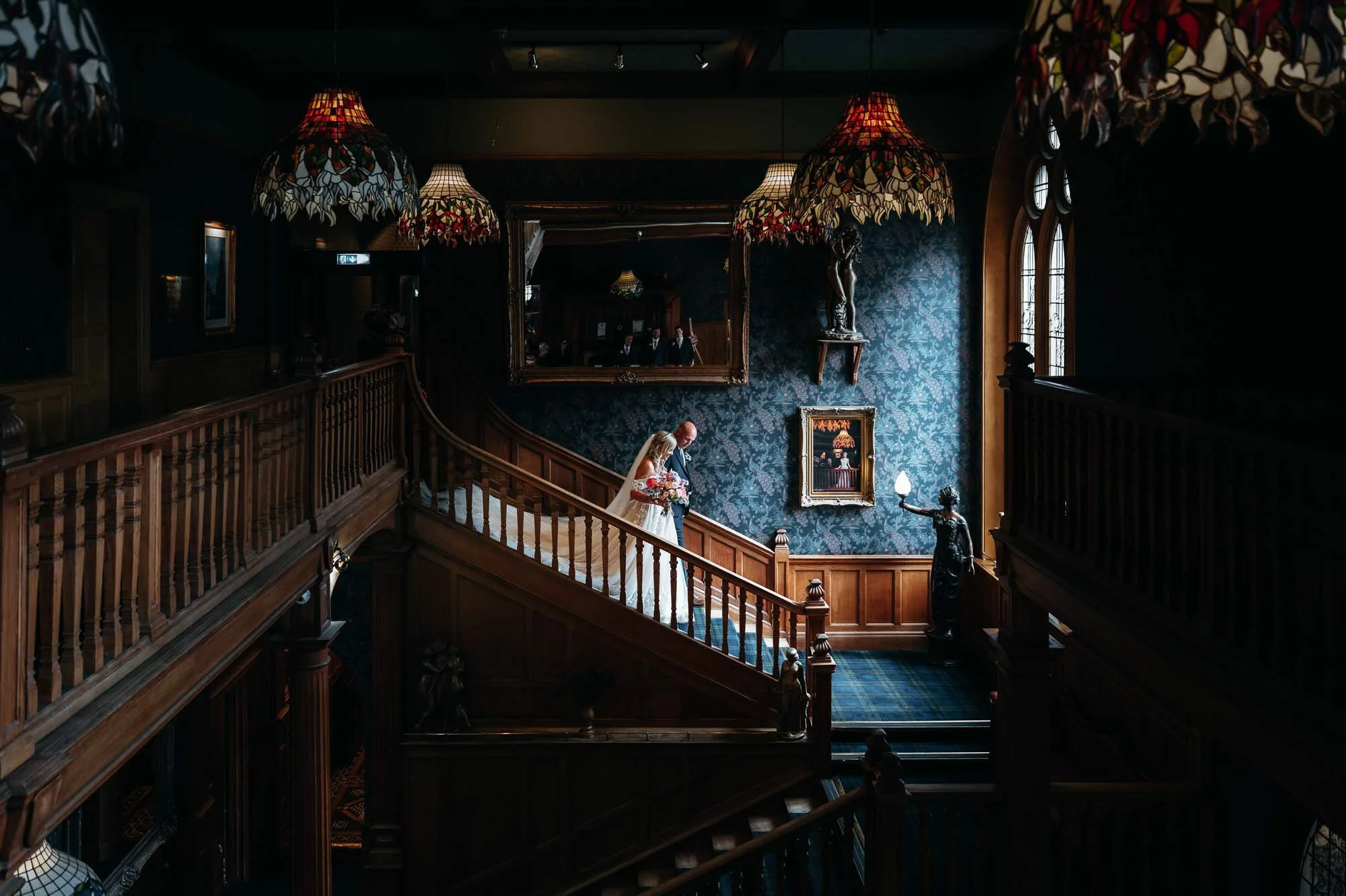 Interior of a dimly lit vintage hotel or mansion with dark blue patterned wallpaper, wooden staircases, ornate lampshades, a large mirror, and classical sculptures.