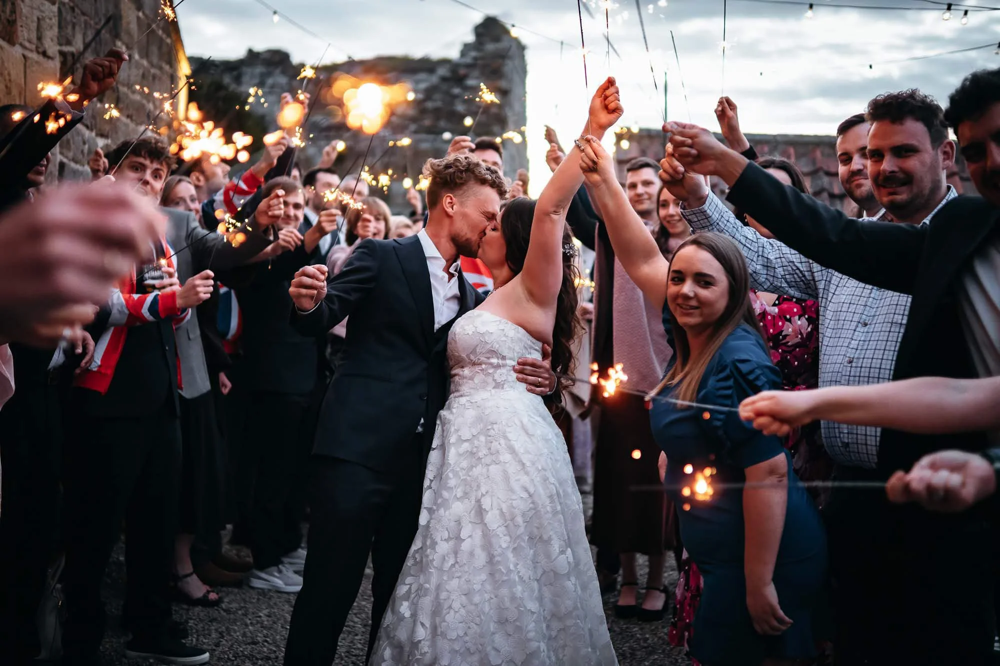 A bride and groom kiss amid a crowd holding sparklers during an outdoor celebration at dusk.