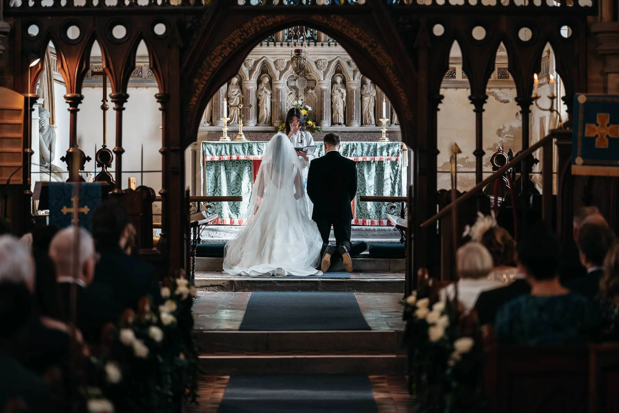 A bride and groom kneeling at the altar during a wedding ceremony inside a church. The altar is decorated with candles and religious statues in the background.