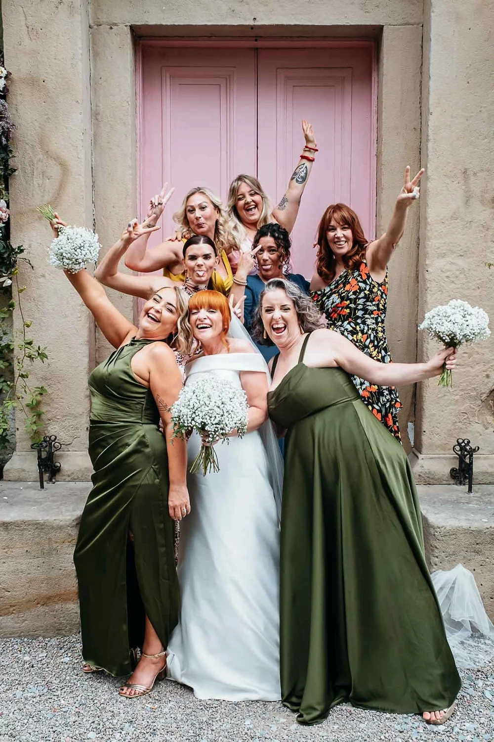 A bride laughing with her bridesmaids in olive green dresses at the pink door of The Croft Hotel