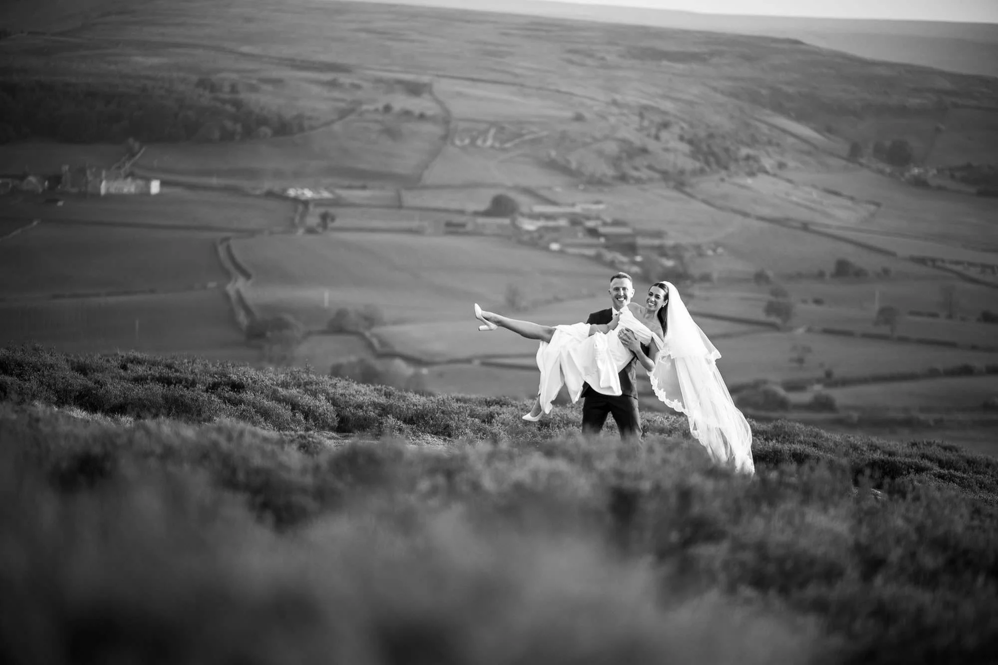 A smiling bride with a veil being carried by a smiling groom across a scenic grassy hill with a rural landscape in the background.