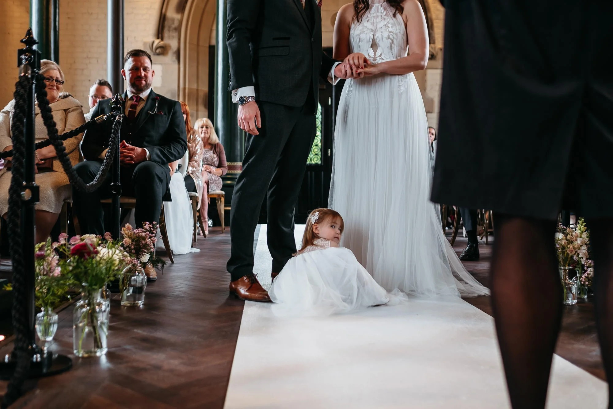 A young girl in a white dress sitting on the wedding aisle during a wedding ceremony, with the bride and groom holding hands at the altar. Guests are seated in the background, observing the ceremony.