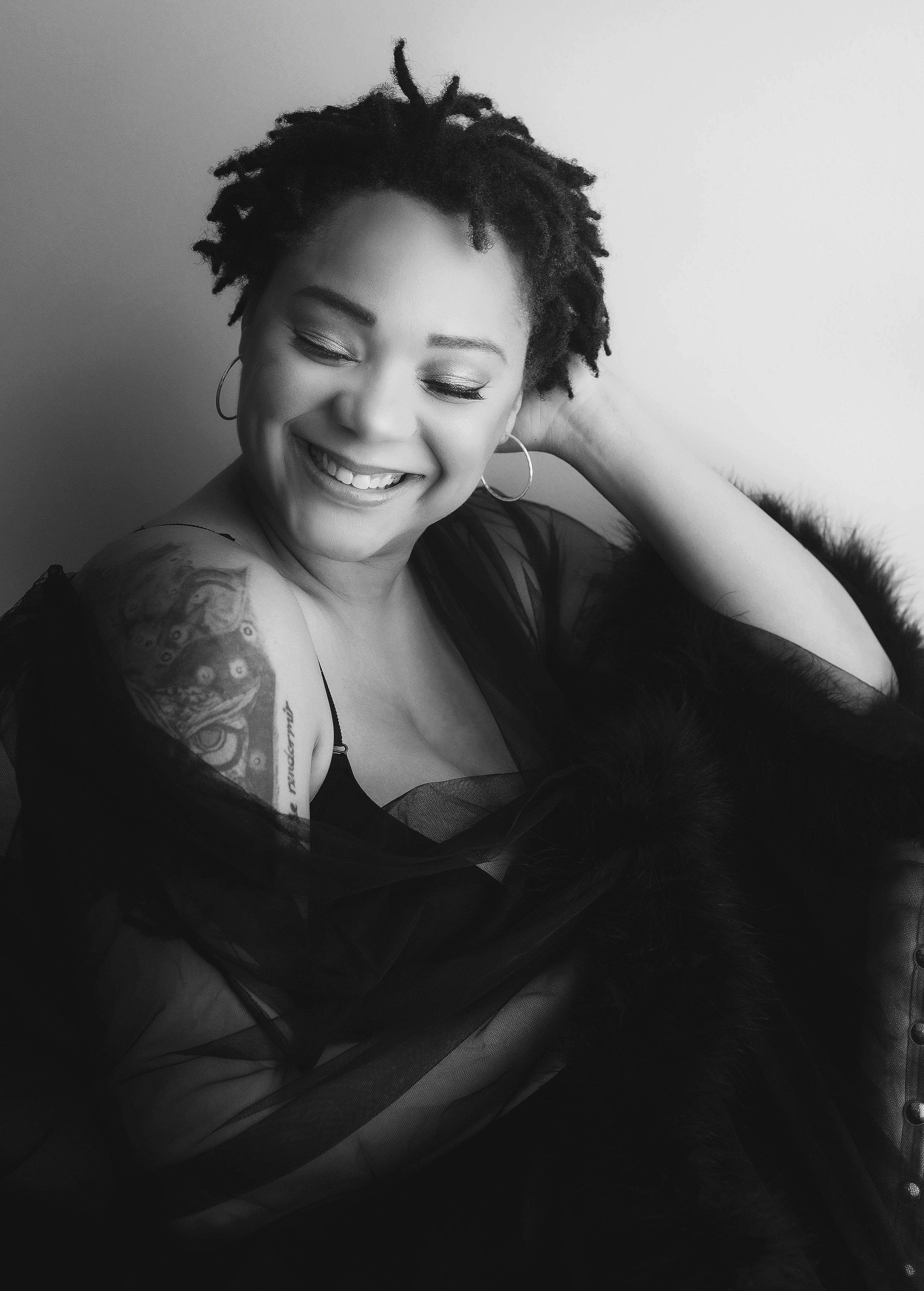 A smiling woman with short, curly hair, wearing hoop earrings and a sheer black top, sitting with her eyes closed and leaning slightly to the side.