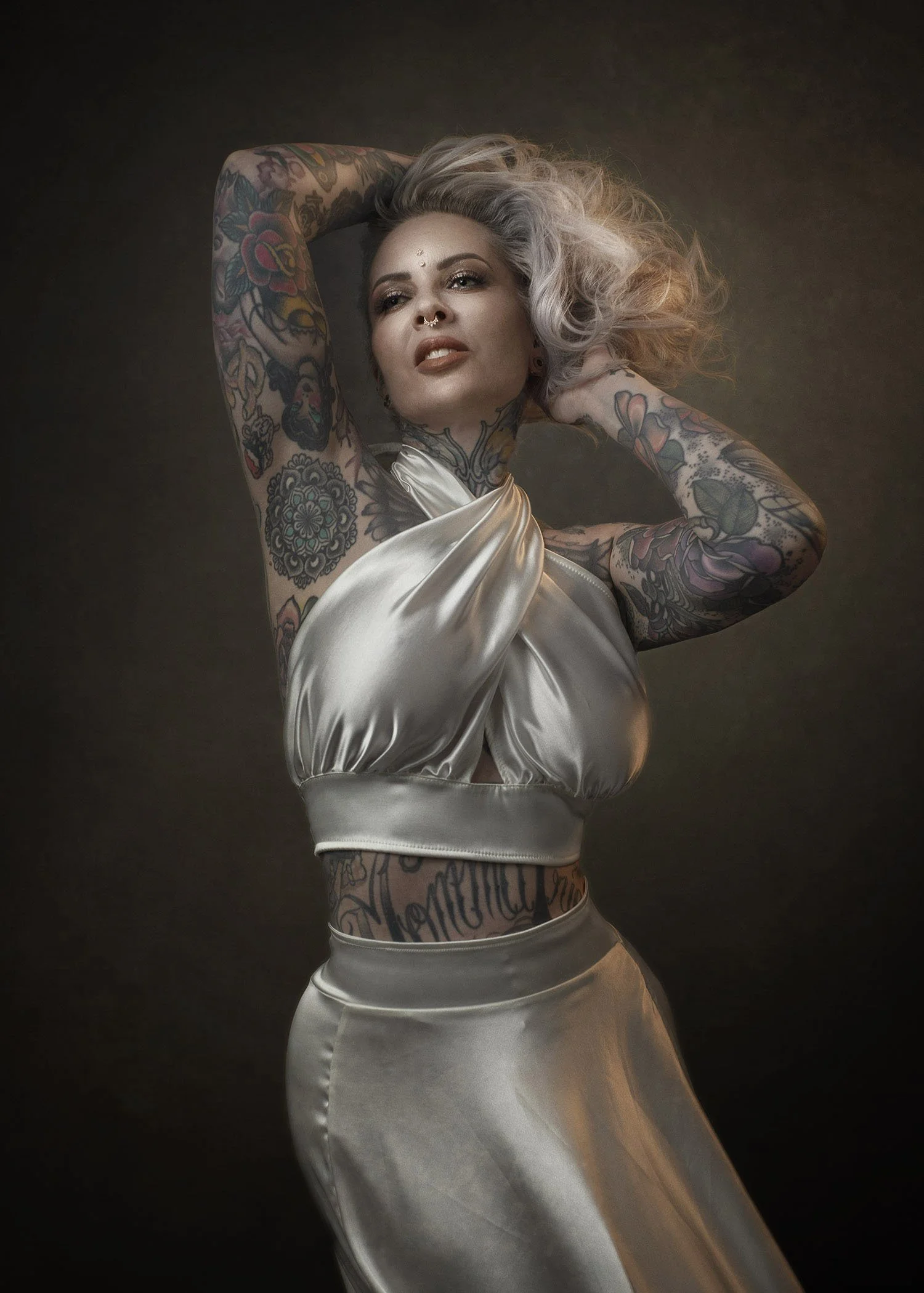 Stylish woman with platinum blonde hair and tattoos wearing a shiny satin silver top and matching skirt posing with arms raised behind head against a dark background.