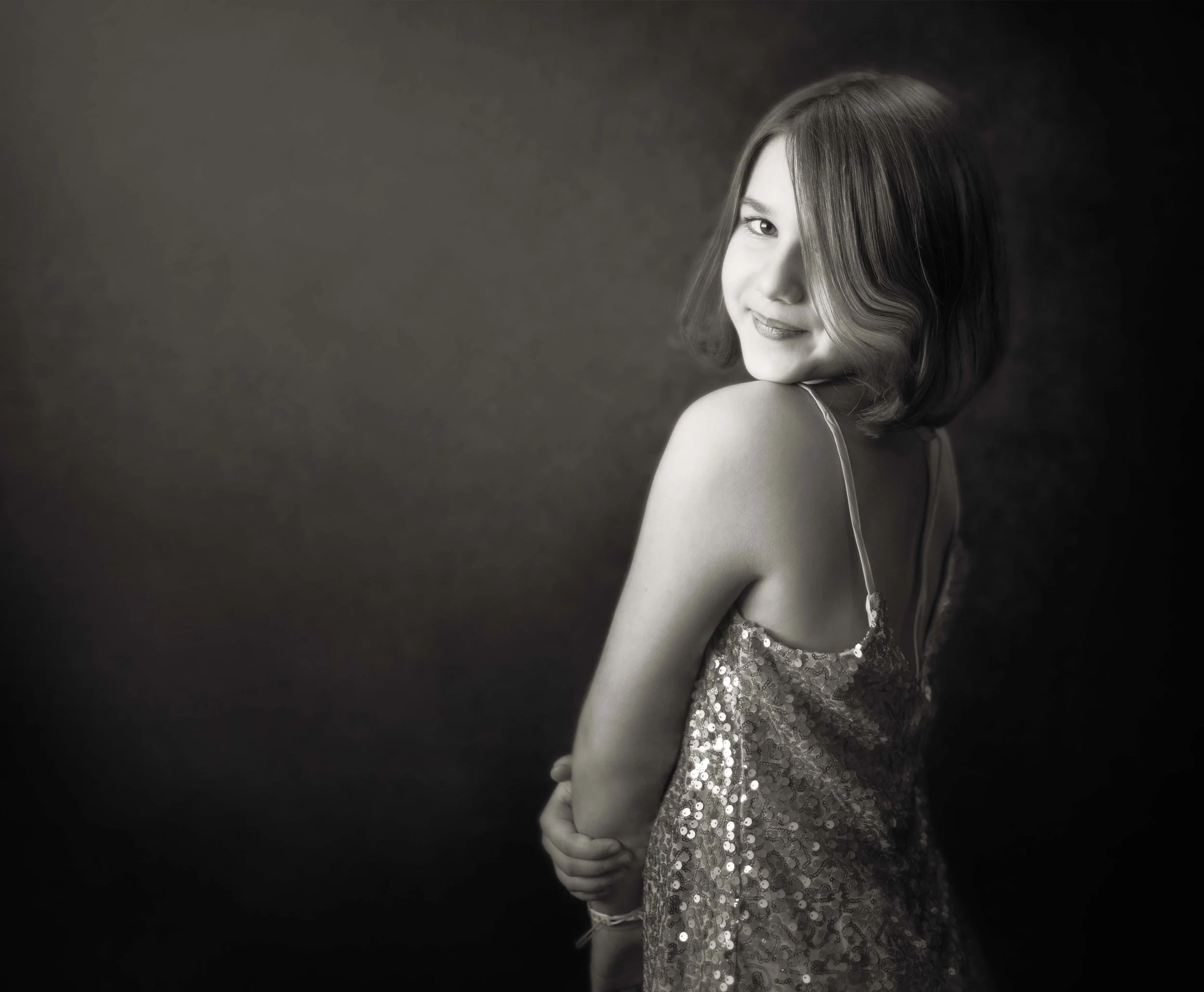 A young woman with shoulder-length hair, smiling while looking over her shoulder, wearing a sparkly dress, in a black and white portrait.