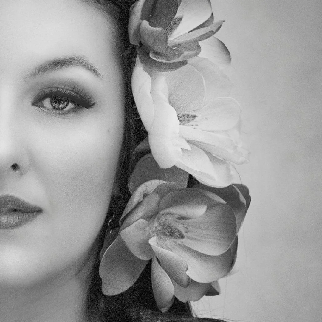 Black and white photo of a woman with makeup, with part of her face visible and a large flower near her head.