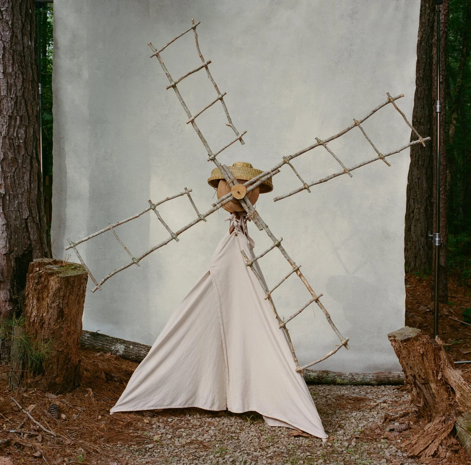 A decorative windmill made of sticks, with a fabric cover resembling a skirt, in a wooded outdoor setting.